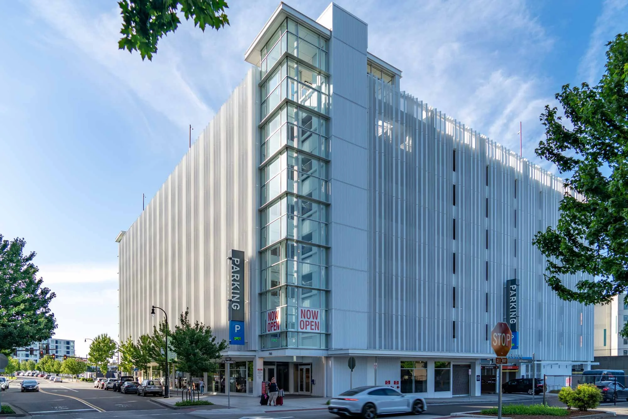 A modern multi-story parking garage with a white exterior, glass windows, and signage indicating it's now open. The street in front has cars and pedestrians, with trees lining the area and a clear blue sky overhead.