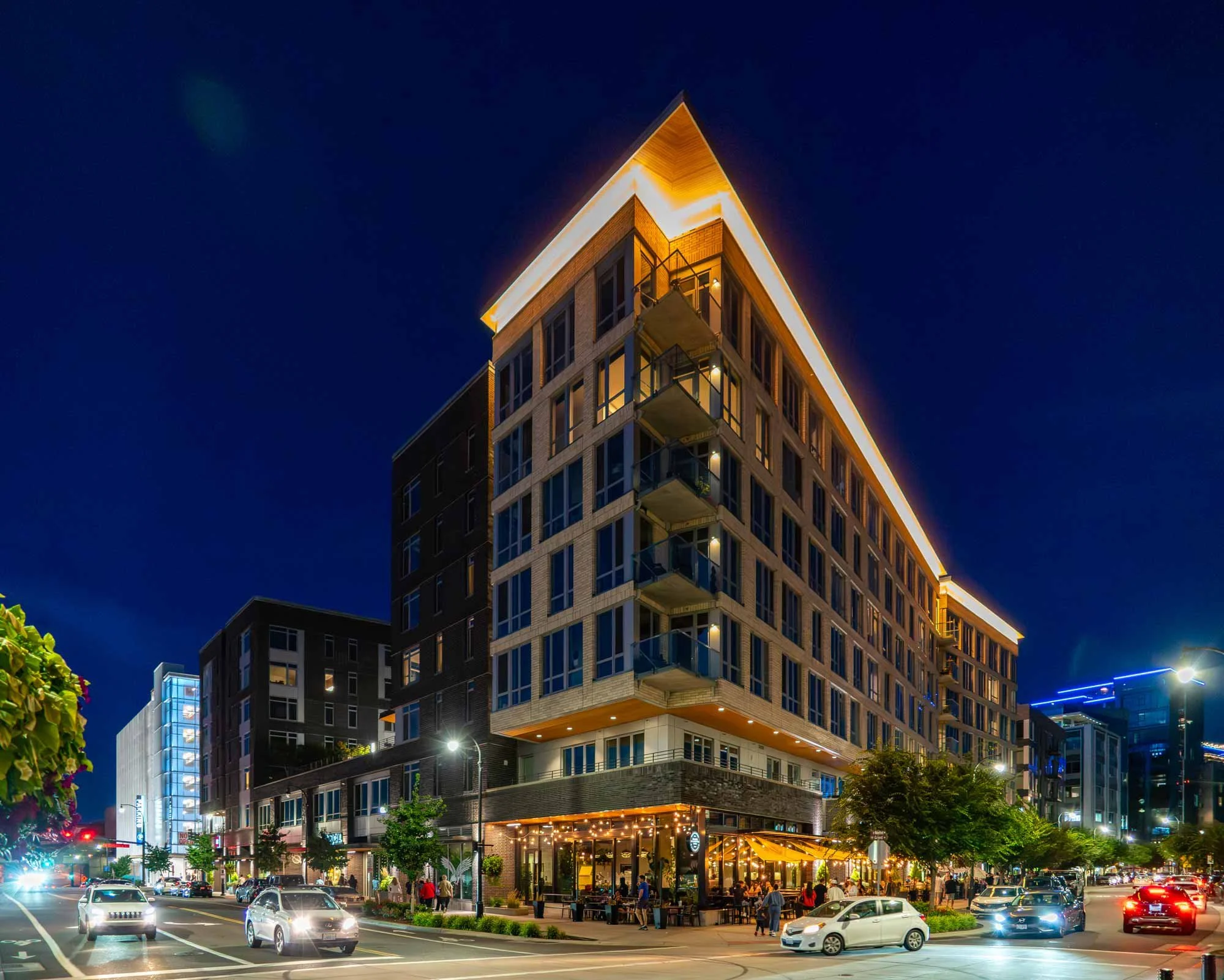 Night view of a modern multi-story building with illuminated roof and balconies, bustling street with cars and people, and trees outside.
