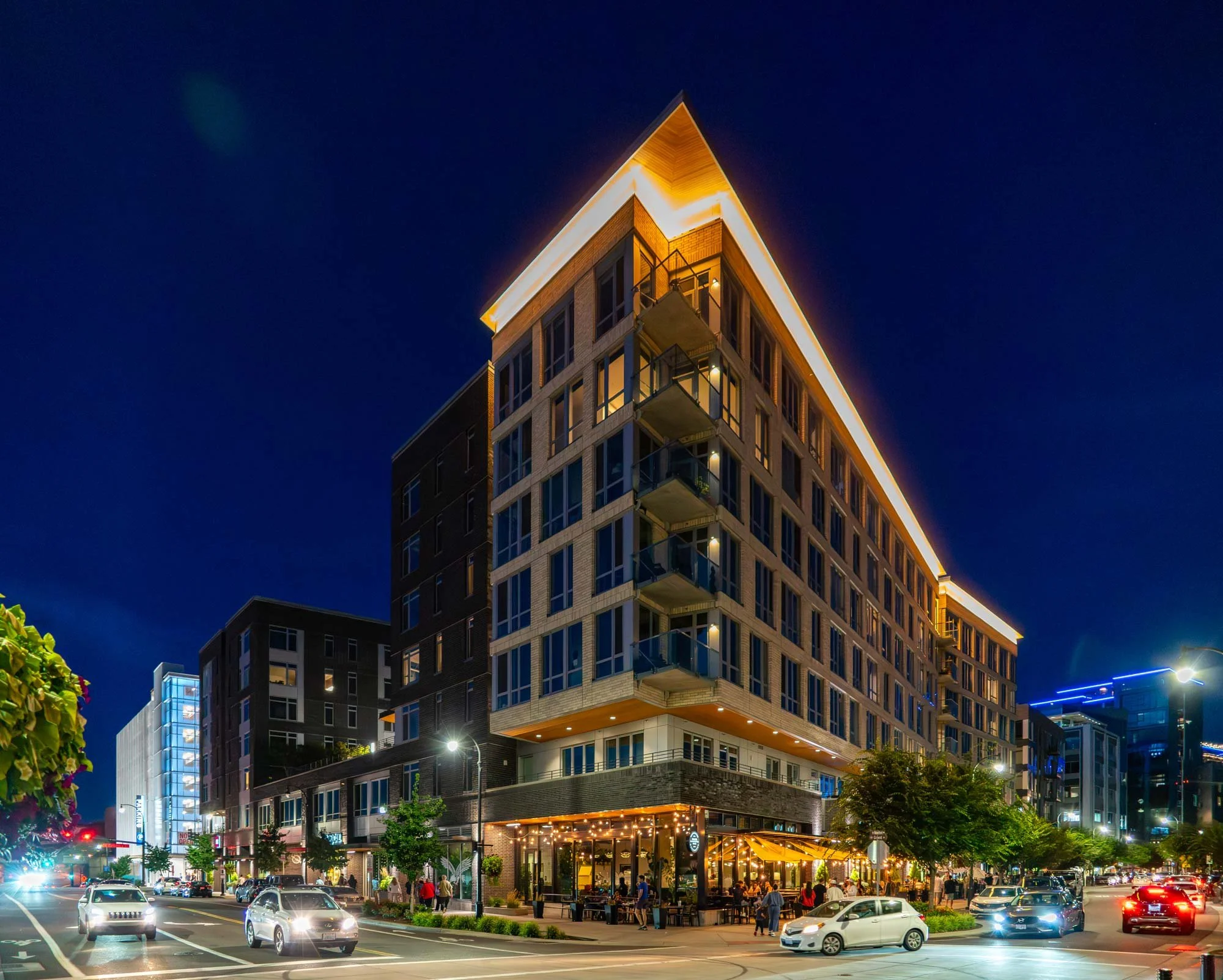 Night view of a modern, lit-up multi-story building with street activity, cars, and pedestrians in an urban area.