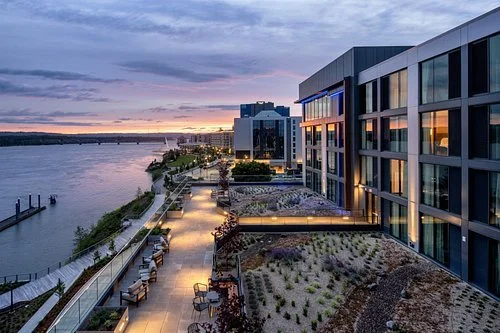 View of a modern urban waterfront with a boardwalk, outdoor seating, and high-rise buildings during sunset.