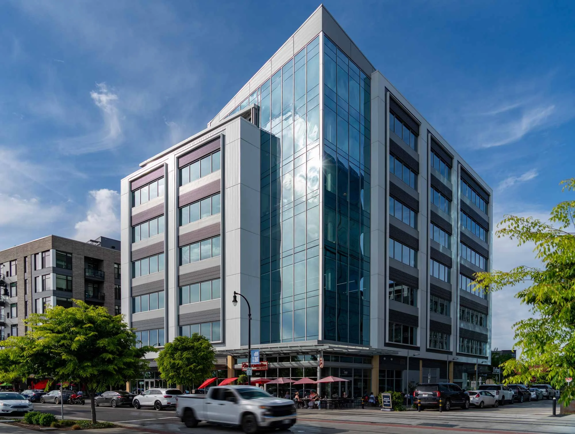 Modern office building with glass windows reflects the sky and clouds, with trees, parked cars, and a street cafe with tables and umbrellas in the foreground.