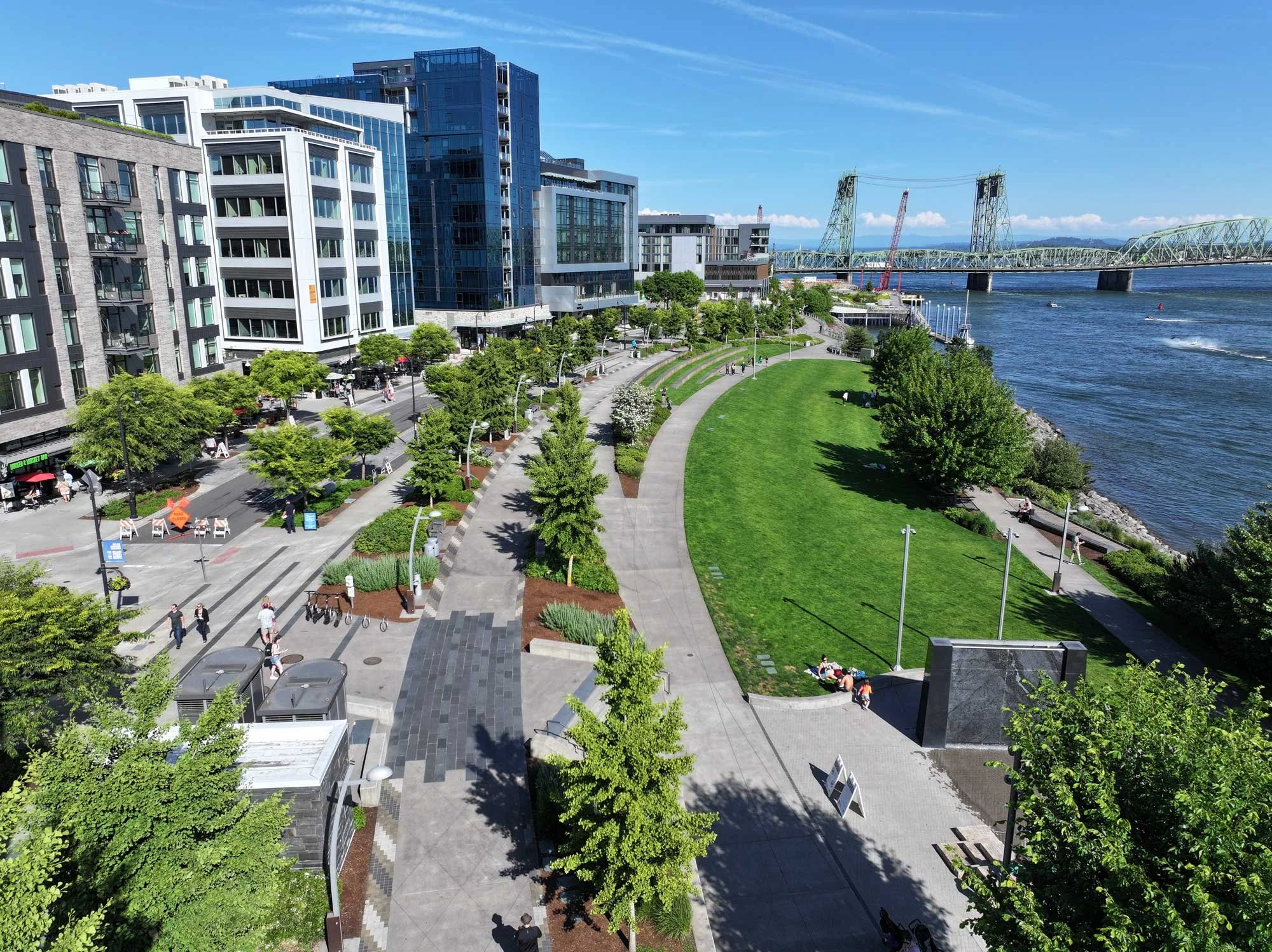 A city riverside park with pathways, green grass, trees, and modern multi-story buildings, with a bridge and water in the background under a clear blue sky.