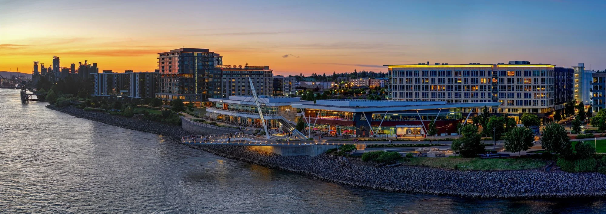 Cityscape at sunset with modern buildings along a river, featuring a waterfront park and illuminated structures.