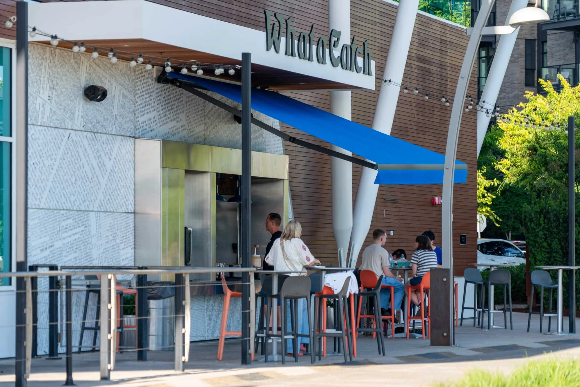 Outdoor seating area at Wild Earth, with people sitting at tables, and a modern building with wood and concrete exterior in the background.