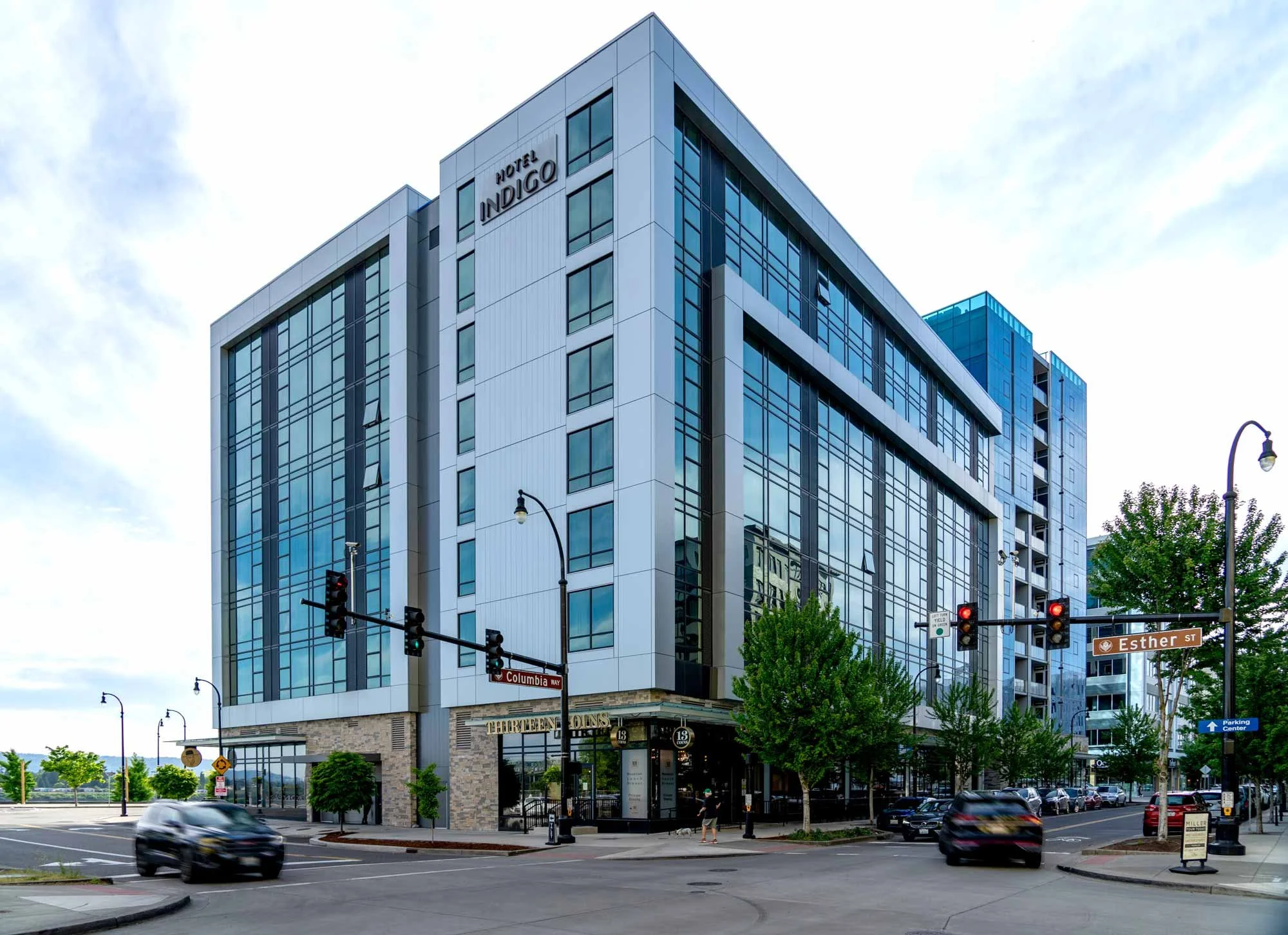 A modern multi-story hotel building called Hotel Indigo with large glass windows, located at the intersection of Columbia Way and Esther Street. There are street signs, traffic lights, and some cars and pedestrians visible in the foreground, with a cloudy sky above.
