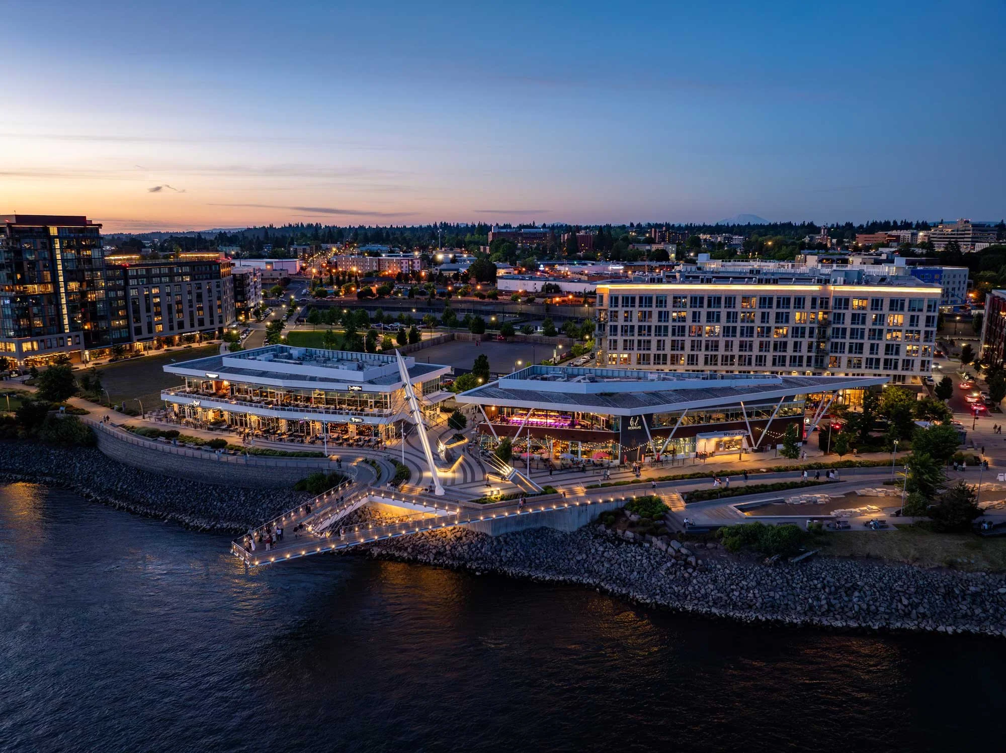 A cityscape at dusk featuring modern buildings along a waterfront with a illuminated pier and walkway, trees, streetlights, and distant mountains in the background.