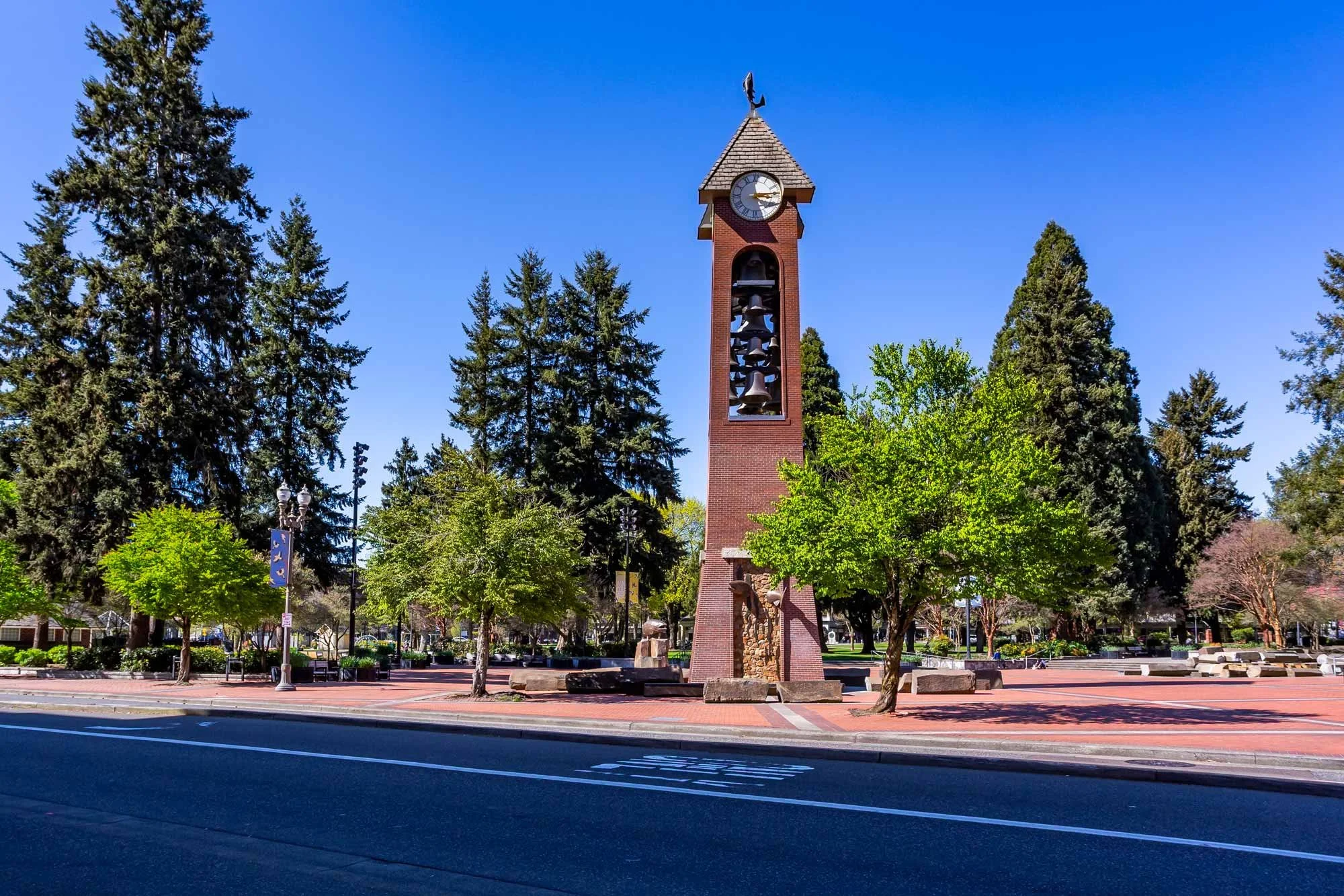 A brick clock tower with a weathervane on top, surrounded by green trees and a park with benches, streetlamps, and banners on a sunny day.