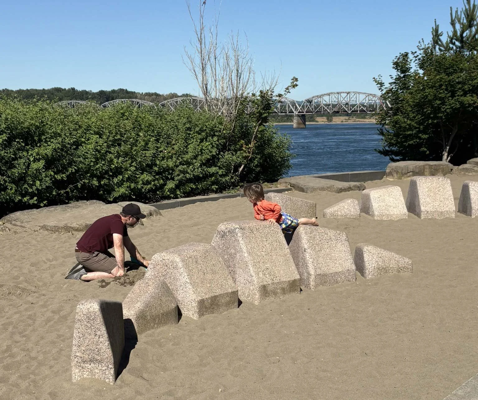 A man and a young boy playing in the sand on a beach, with a row of concrete blocks nearby and lush green bushes behind them. In the background, there's a river with a bridge crossing over it under a clear blue sky.