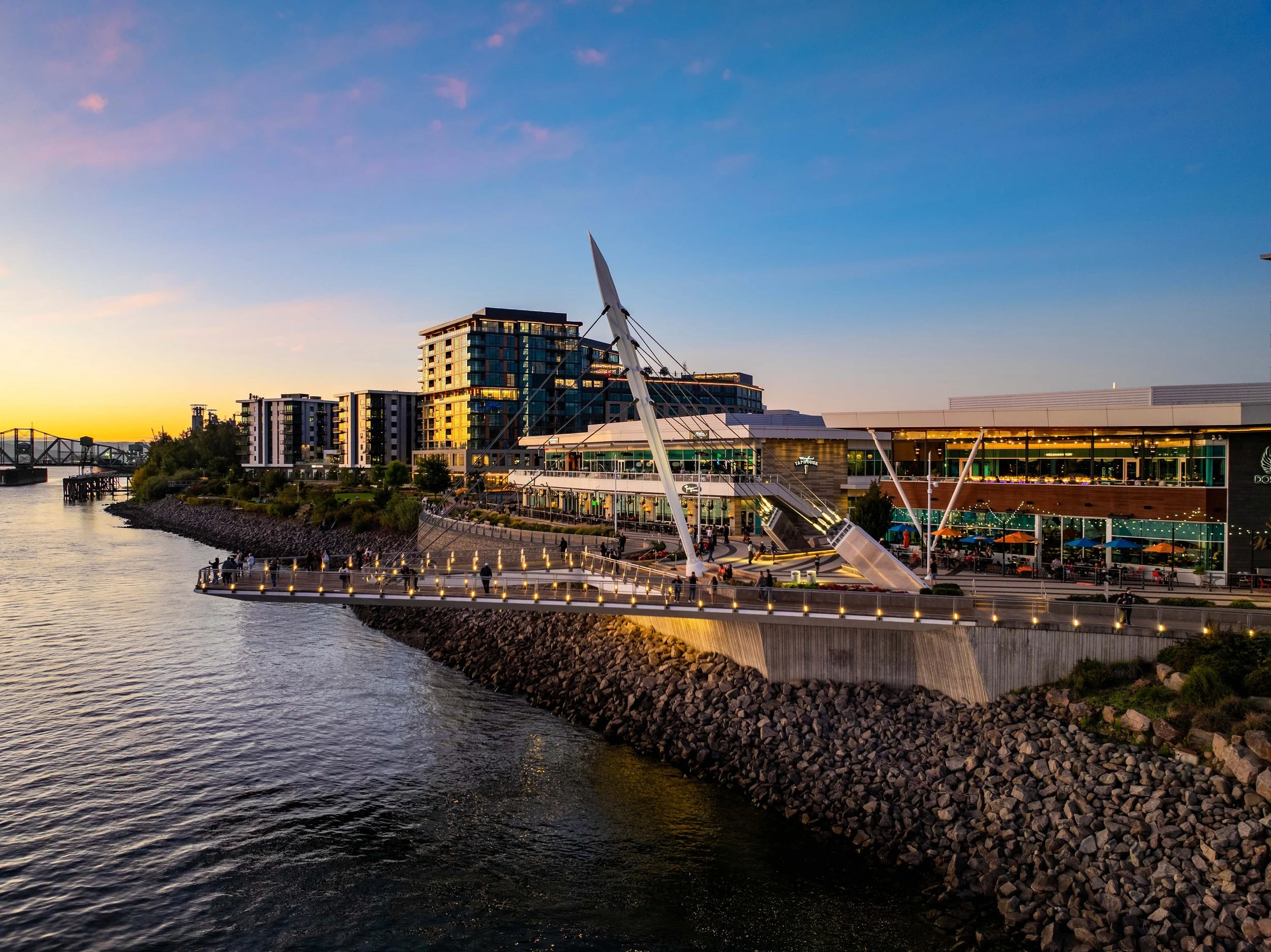 A waterfront scene during sunset featuring modern buildings and a bridge, with a pier and people walking along the shore.