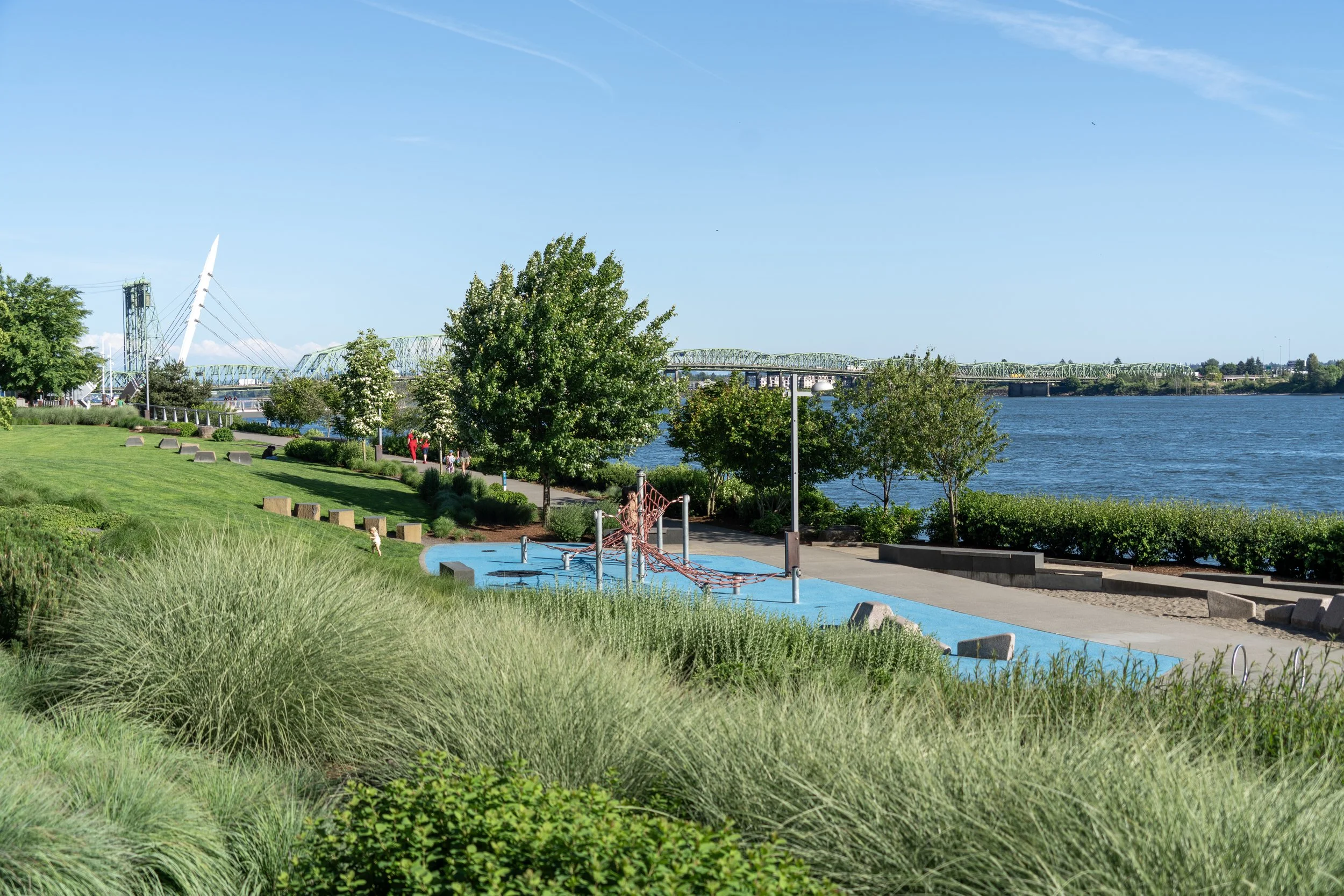 Scenic Waterfront Vancouver park next to the Columbia River with a bridge in the background. The park has green grass, trees, bushes, and a playground with children playing, under a bright blue sky.
