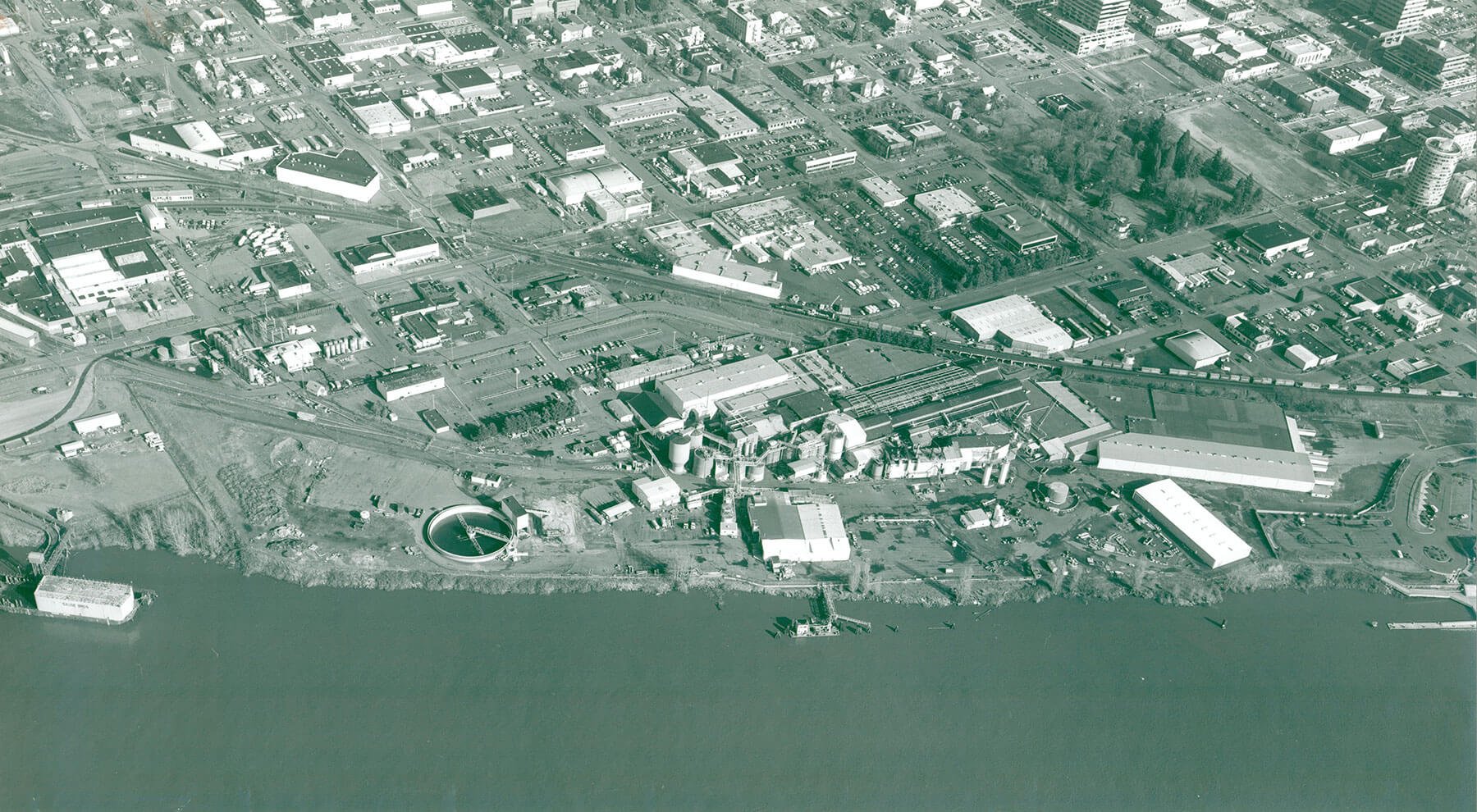 Aerial view of a cityscape next to a body of water, featuring industrial and commercial buildings, roads, railways, and a circular water treatment facility.