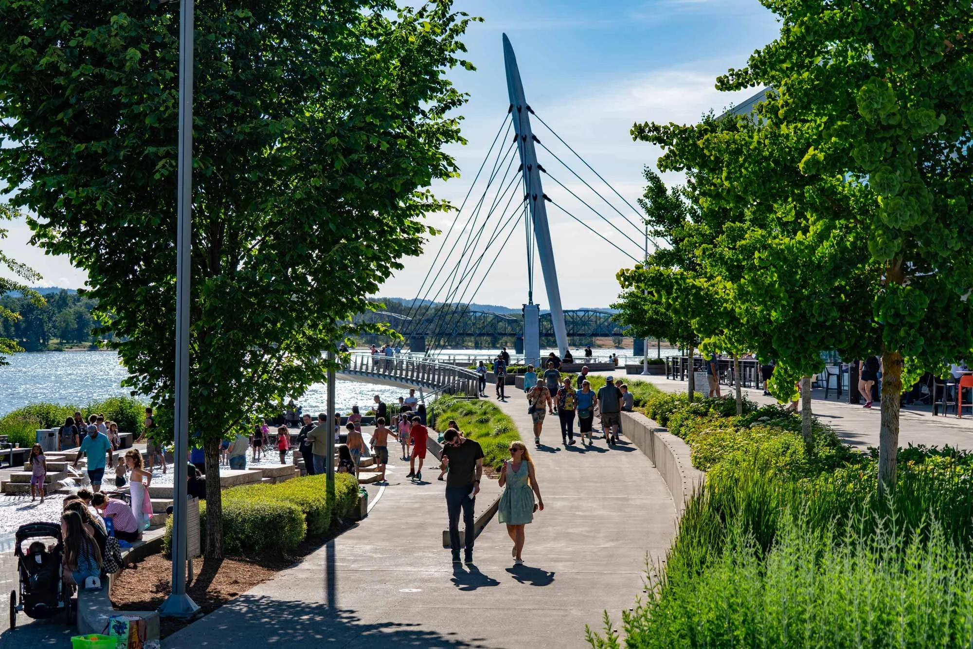 A busy riverside park with people walking and socializing on a sunny day. Trees line the walkway, and a modern bridge and pier are visible in the background.