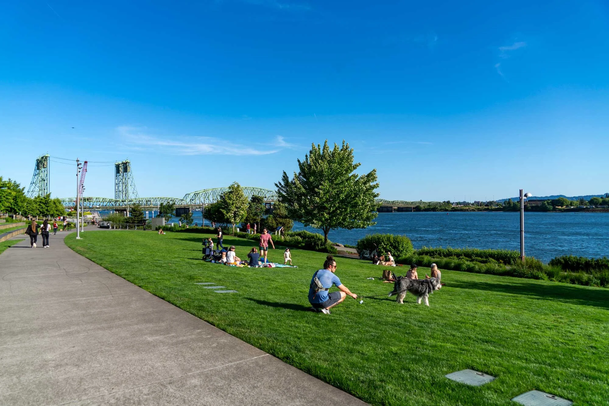 People relaxing and walking in a park alongside a river, with trees, a bridge, and a clear blue sky in the background.