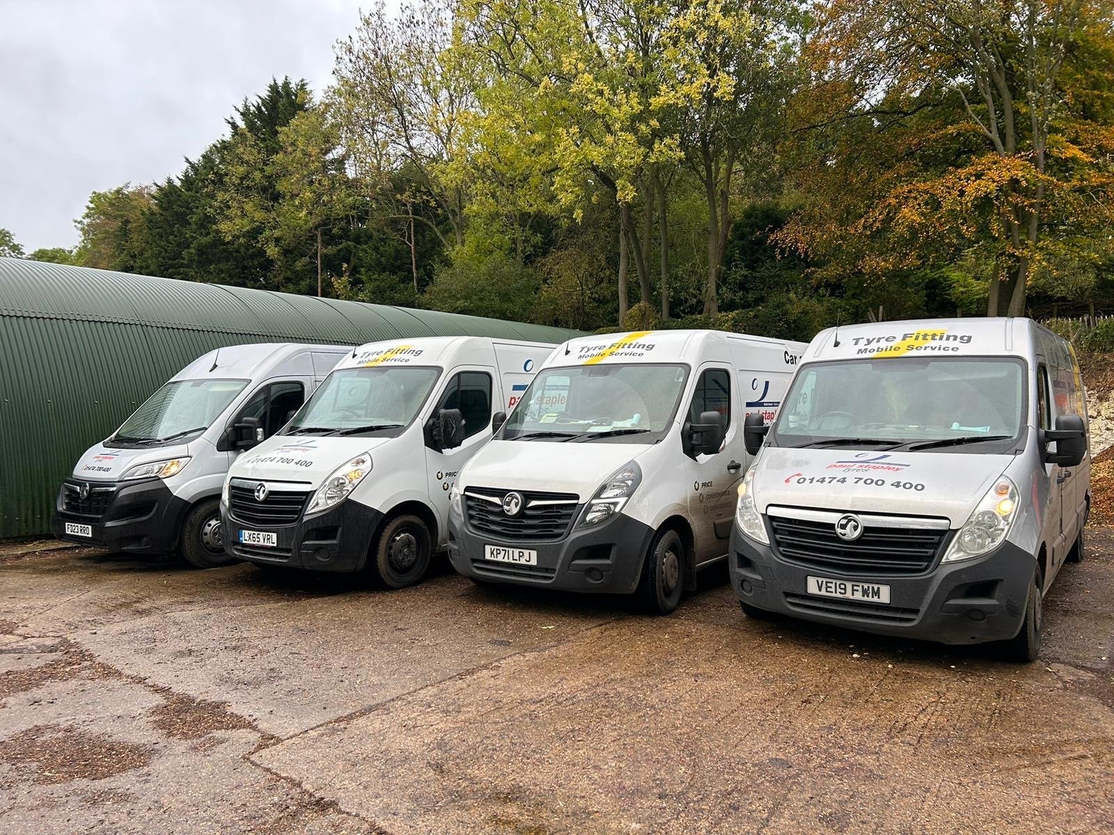 Four white commercial vans parked on a gravel lot, with green trees and a curved green shed in the background.