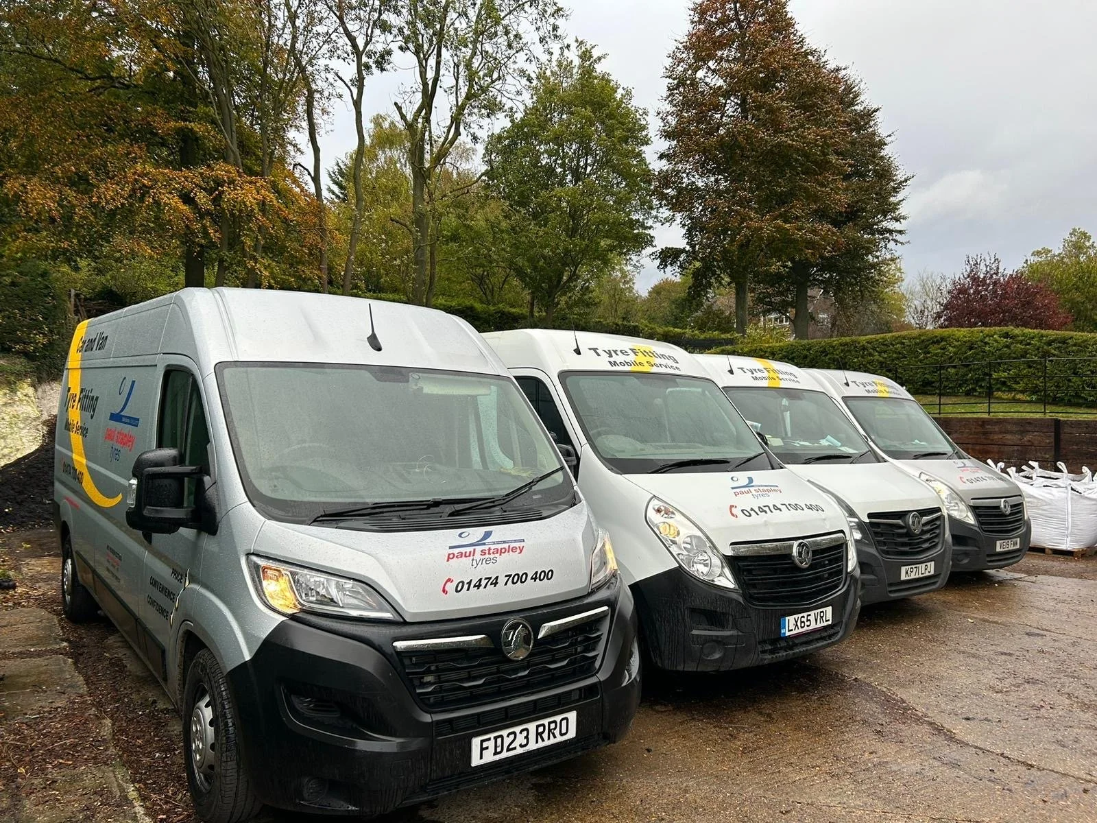 Four company vans parked on a dirt lot, with trees and greenery in the background, overcast sky.