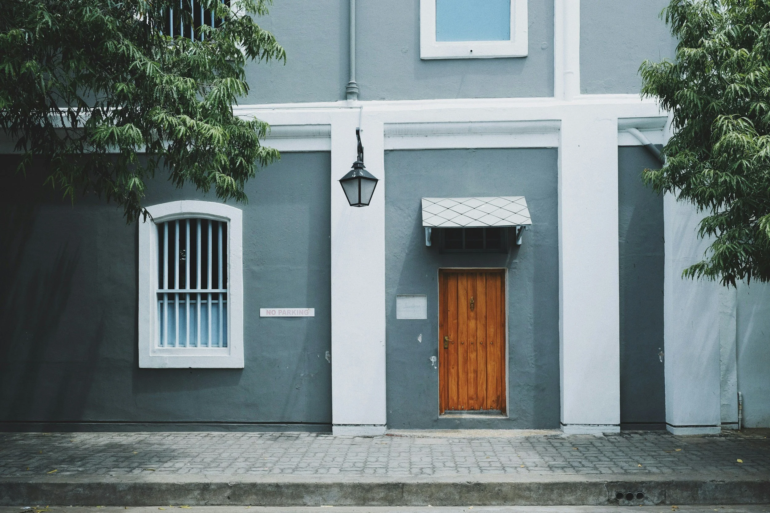 Urban facade with gray walls, white trim, wooden door, window with bars, street lamp, and trees.