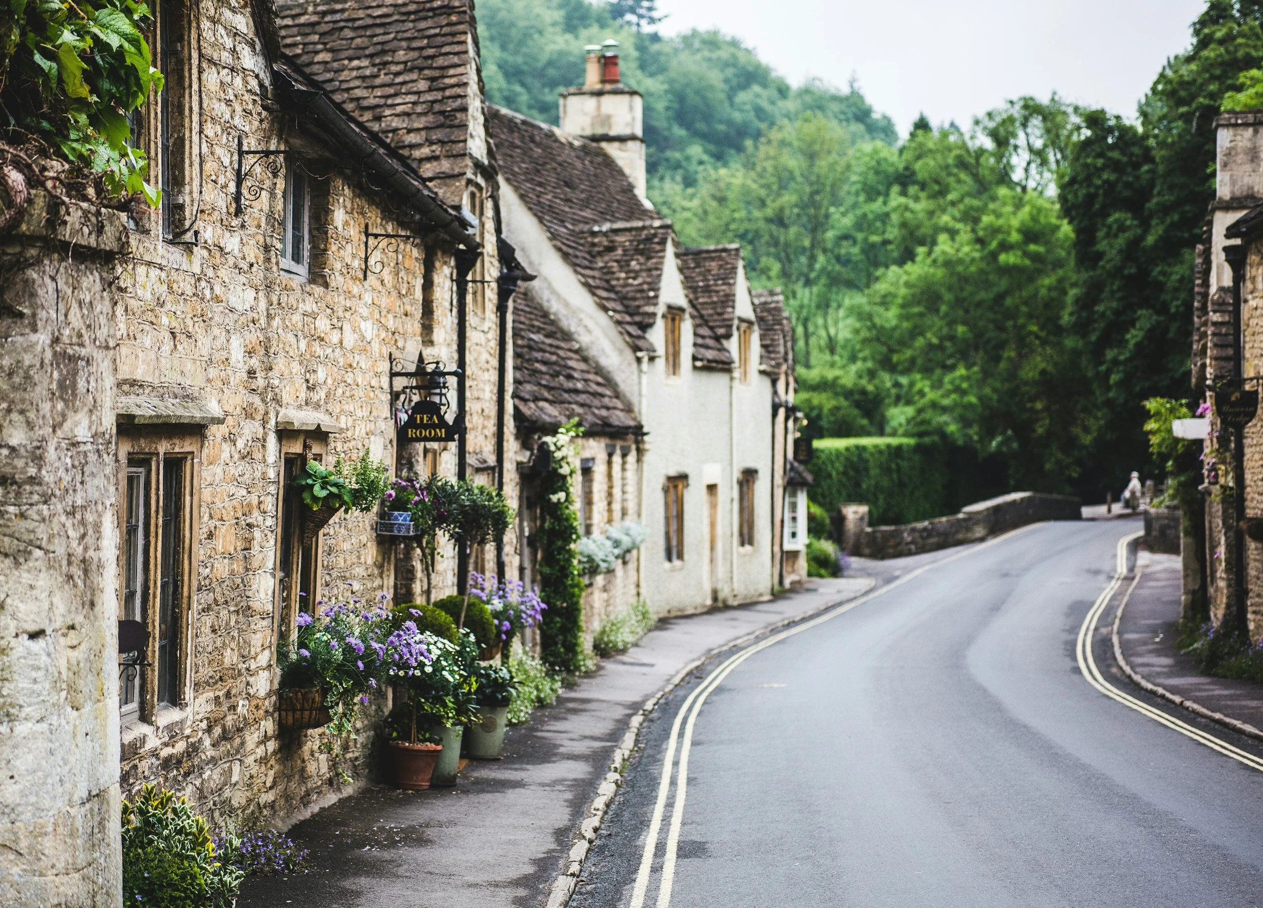 A quaint street lined with stone cottages decorated with flowers and plants, featuring a sign for a tea room, in a countryside setting with lush green trees in the background.