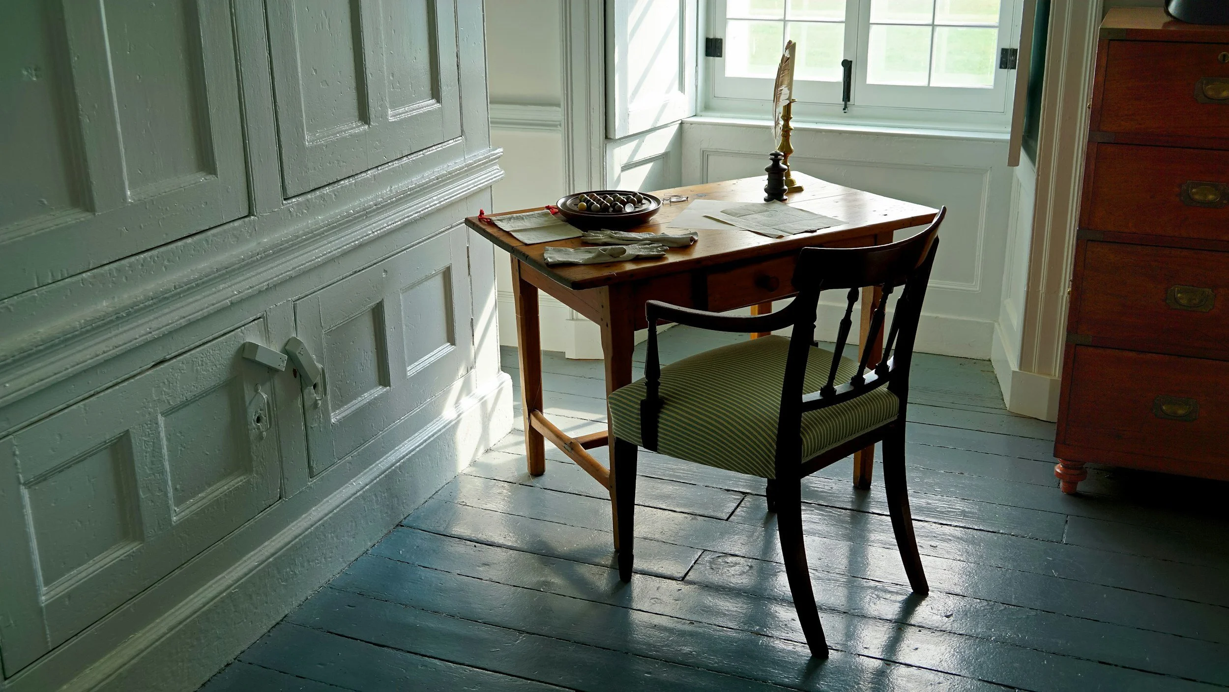 A wooden dining table with a small drawer, placed near a window with sunlight streaming in. The table has a bowl of decorative objects, a couple of papers, and a lamp with a decorative shade. Two chairs are positioned around the table, one with a green and white striped cushion. Next to the table is a tall wooden dresser with brass handles. The room has wooden floorboards and vintage style wall paneling painted in white.