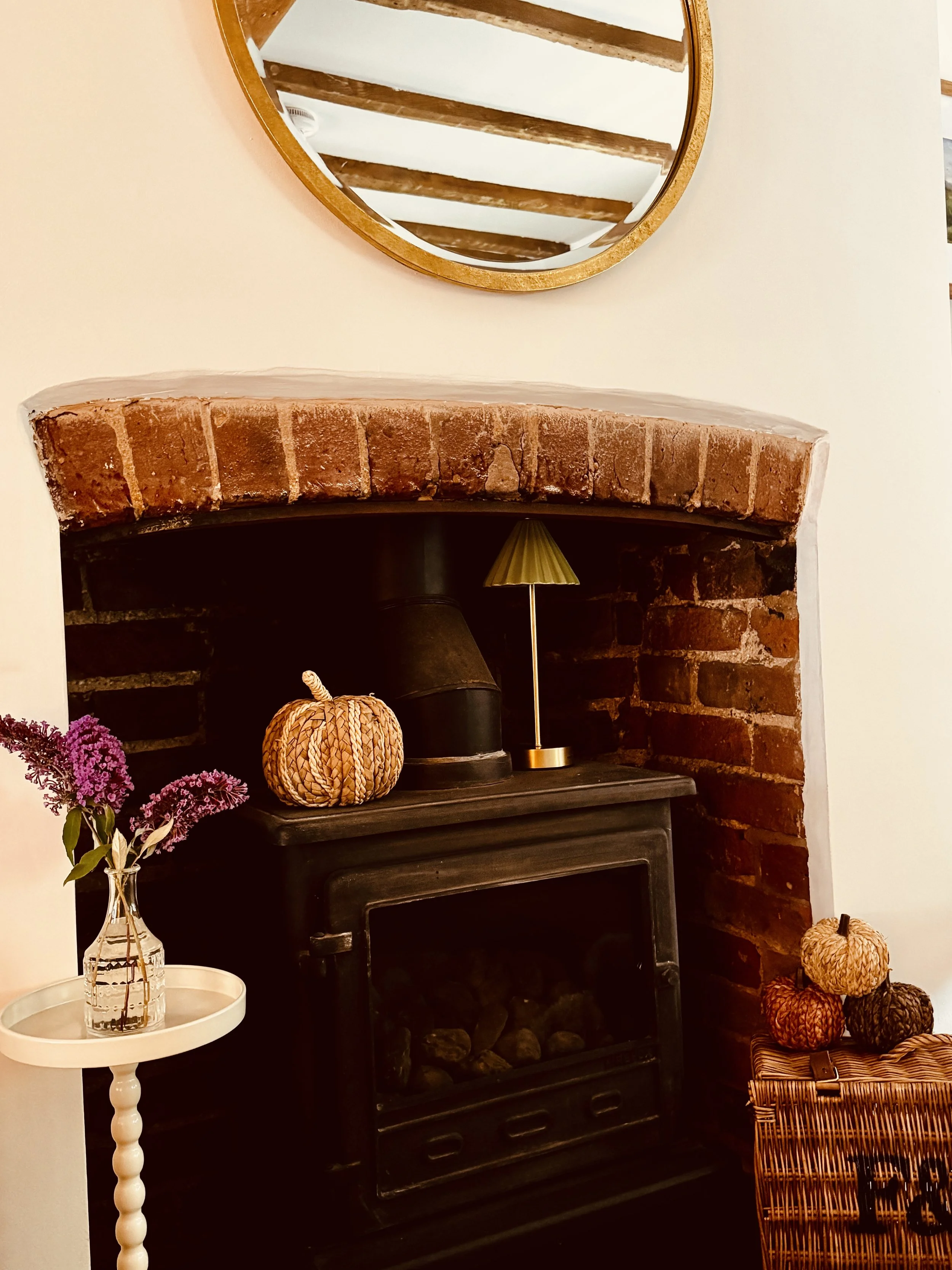 The cottage fireplace and wood-burning stove, decorated with a wicker pumpkin, lamp and flowers.