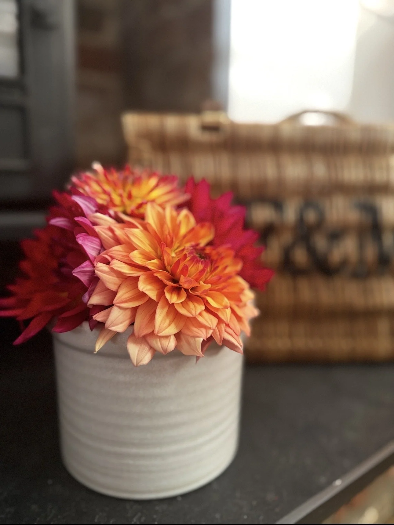 A white ceramic vase with colorful orange, pink, and yellow dahlias, placed on a black surface with a blurred background of furniture and house decor.