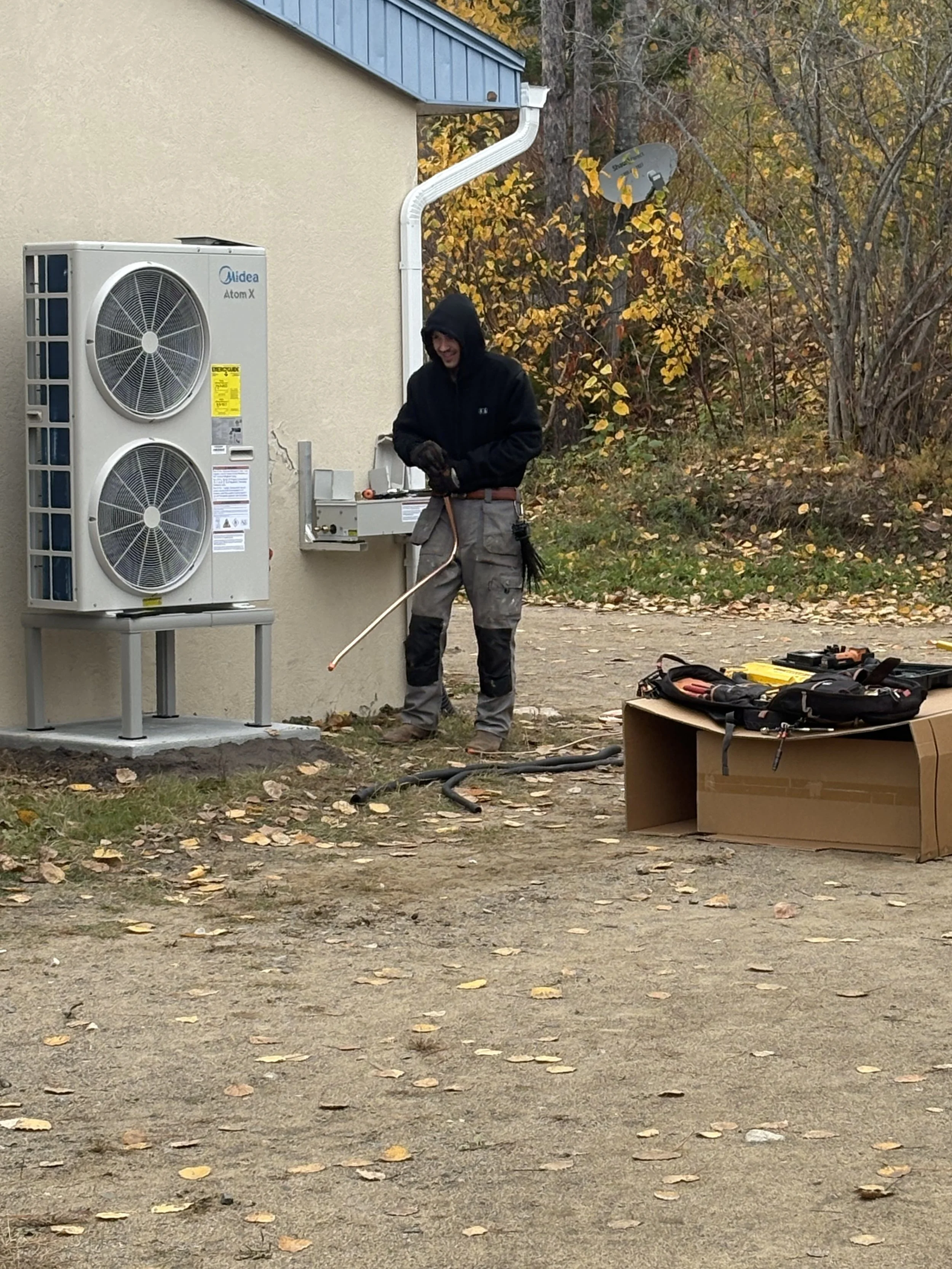 Un technicien effectue une maintenance sur une unité de climatisation extérieure, à l'aide d'outils et d'un sac à outils, dans une zone extérieure entourée d'arbres aux feuilles d'automne.