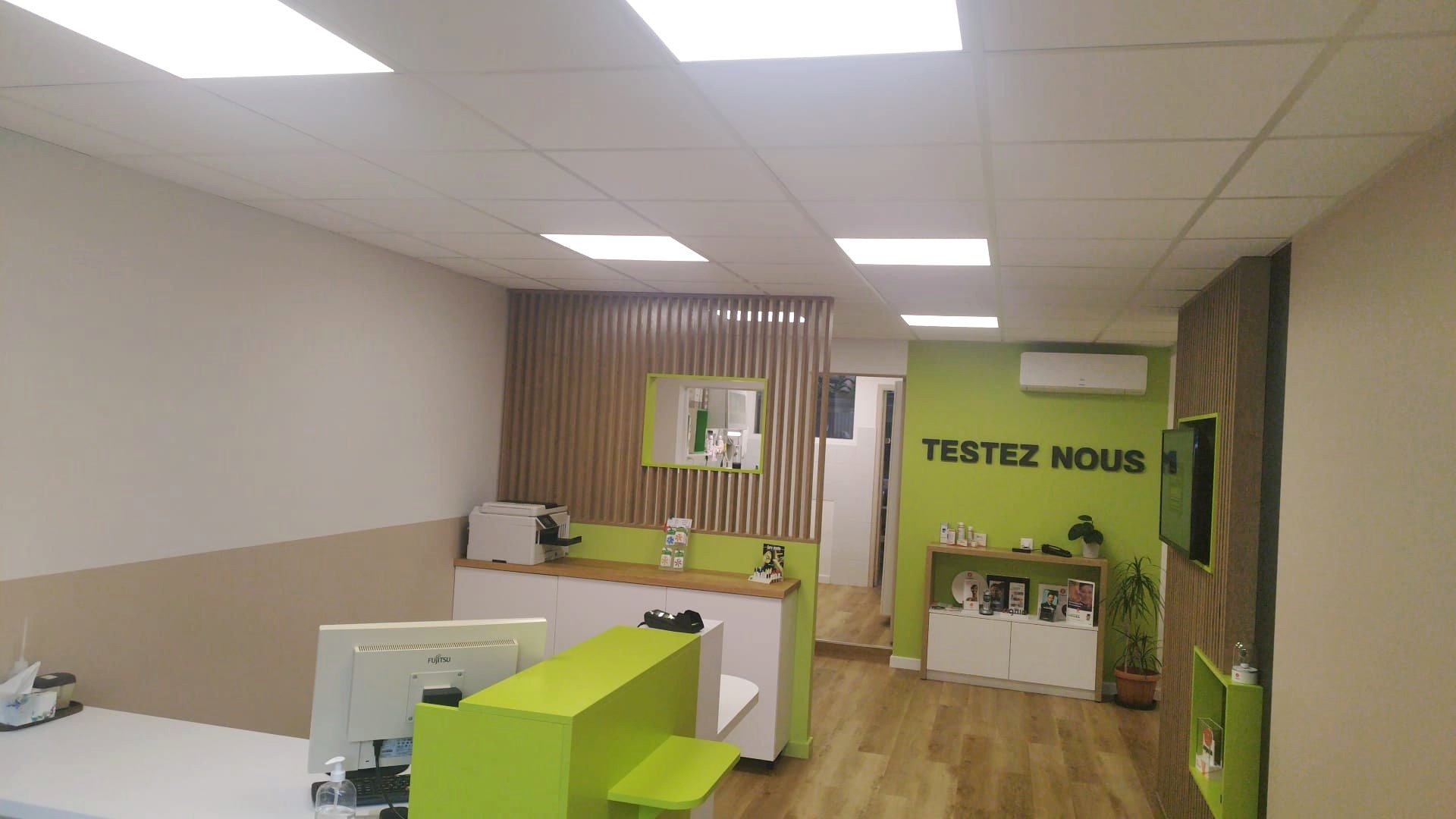 Espace de réception ou salle d'attente avec un mur vert affichant 'TESTEZ NOUS', un bureau avec un ordinateur et une imprimante, et des étagères avec divers produits et brochures, décoration moderne avec bois clair et accents verts.
