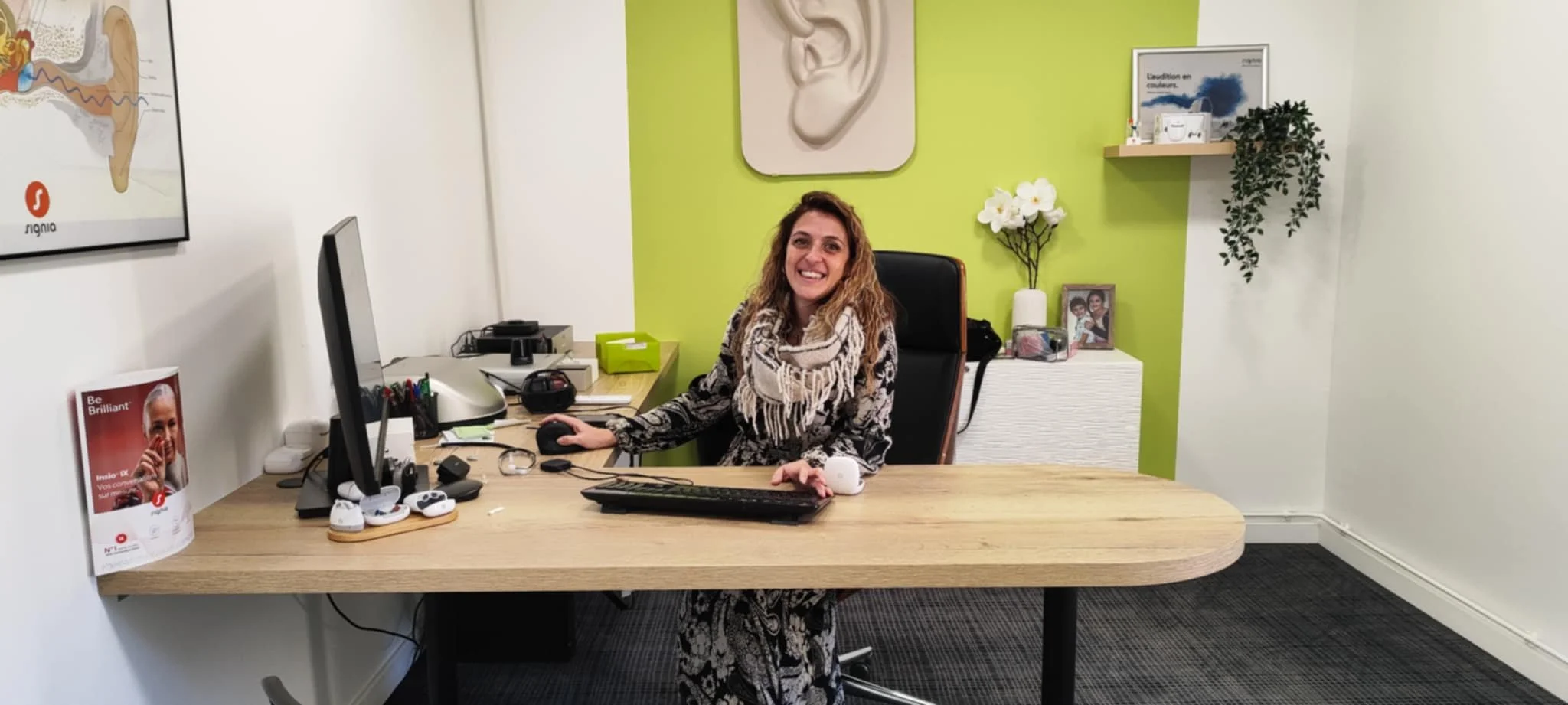 Femme assise à un bureau, souriante, dans un bureau moderne avec mur vert, photo et plantes.