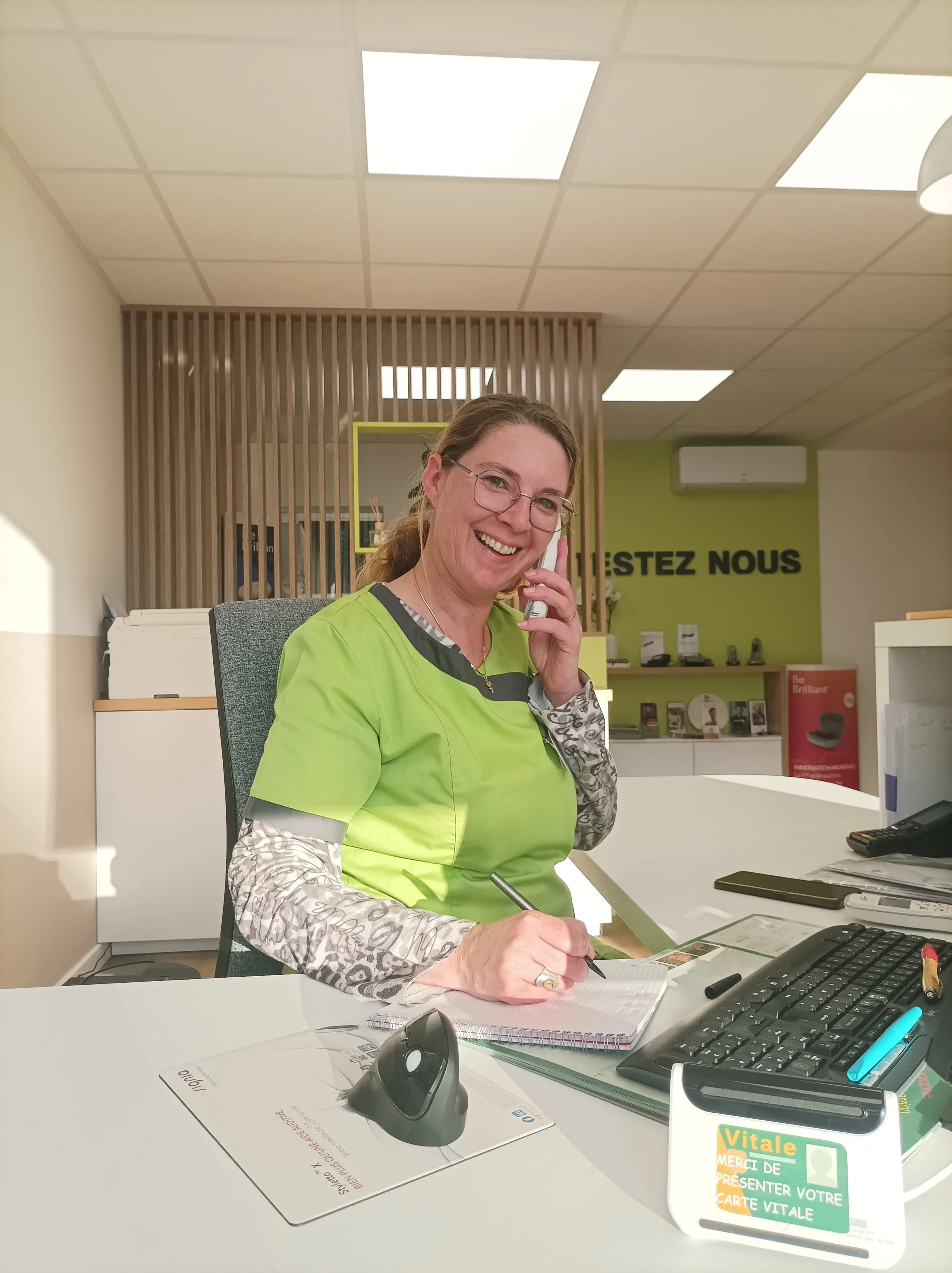 Une femme souriante en uniforme vert assise à un bureau, parlant au téléphone et prenant des notes.