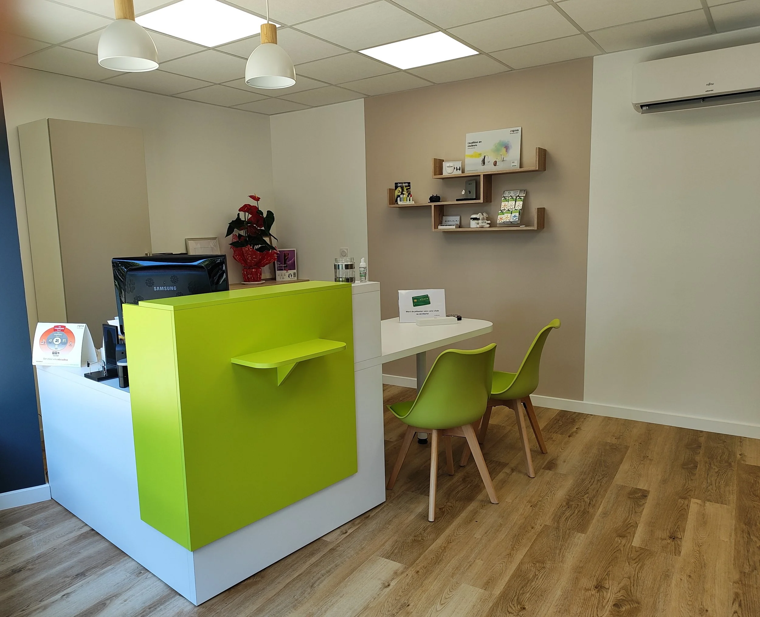 Rendez-vous dans un espace de bureau ou de réception avec un comptoir blanc et vert, deux chaises vertes, un mur avec des étagères en bois et un climatiseur.