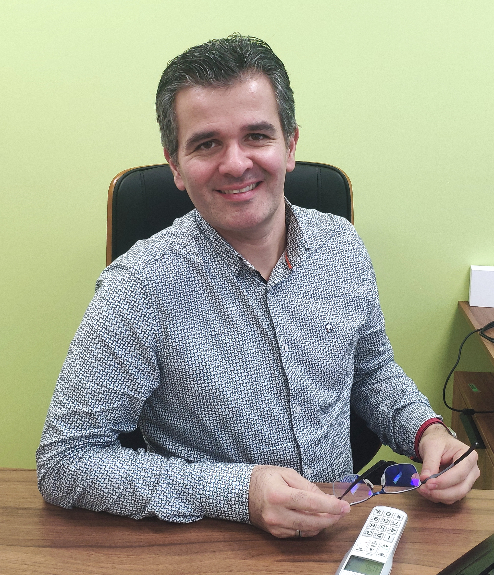 Homme assis à un bureau, tenant des lunettes, avec un fond vert pâle.