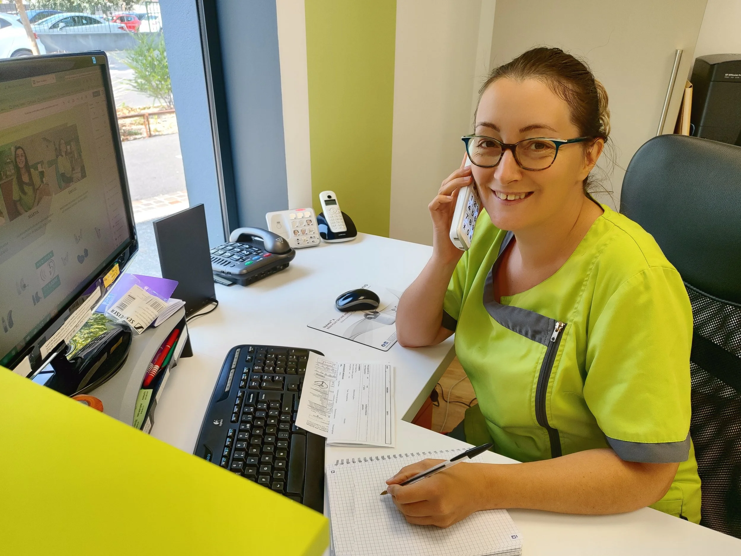Une femme portant une blouse jaune assise à un bureau, parlant au téléphone, avec un ordinateur, un clavier, et des papiers sur le bureau.