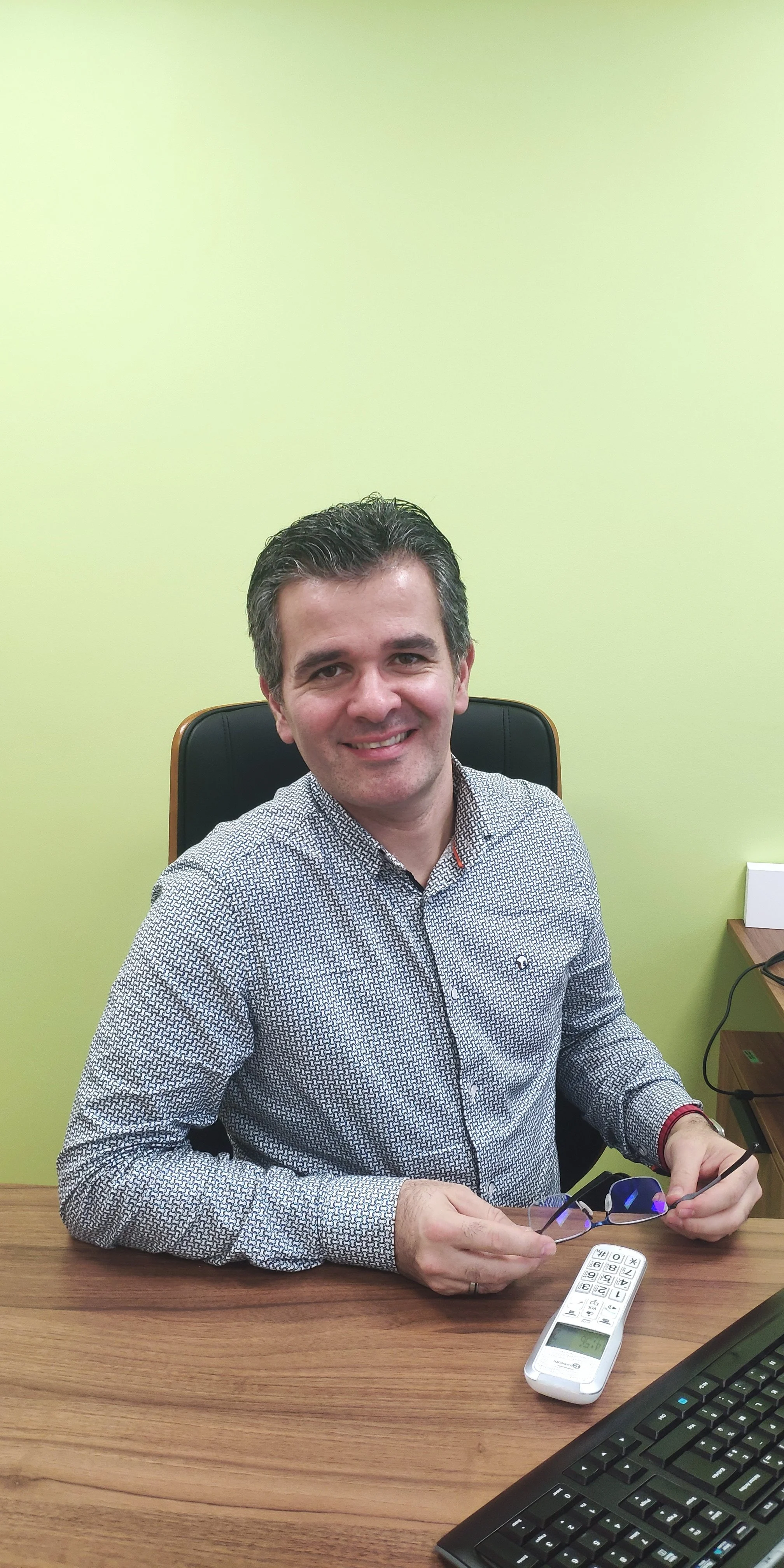 Un homme souriant, assis à un bureau, tenant une paire de lunettes et une télécommande, avec un mur vert en arrière-plan.