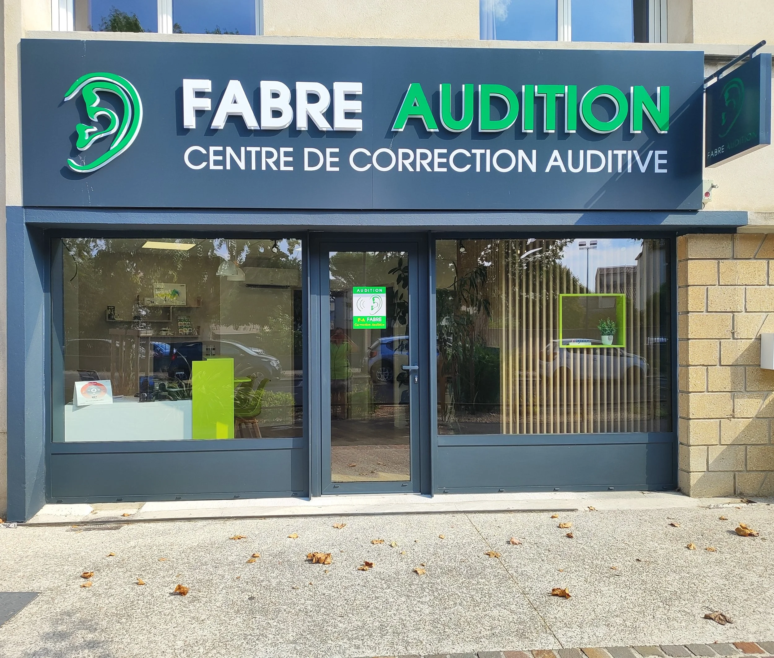 Façade d'un centre de correction auditive nommé FABRE AUDITION, avec un logo d'une oreille, vitres avec affiches et une porte d'entrée.