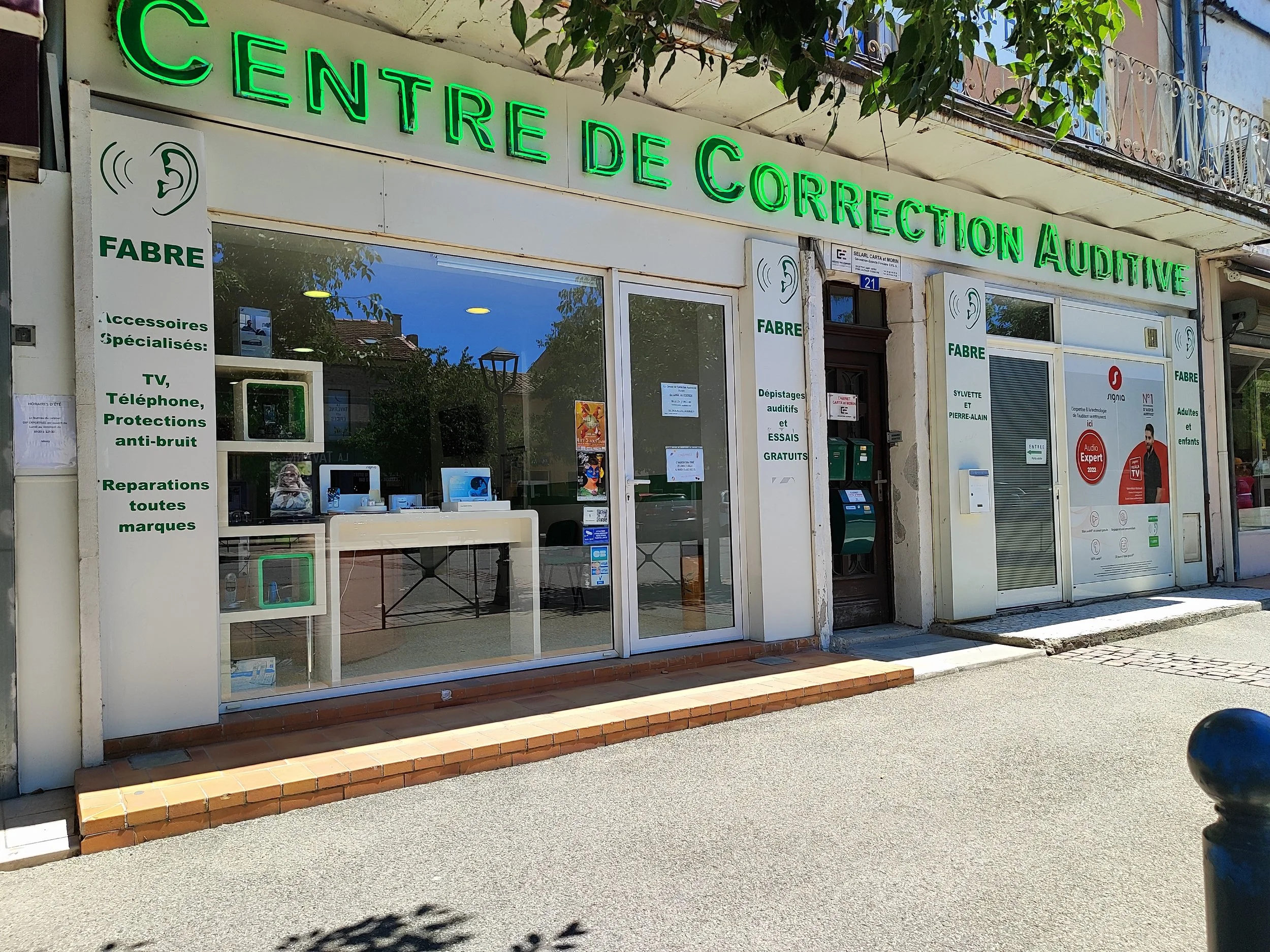 Façade d'un centre d'aide auditive avec des enseignes en néons verts, une vitrine avec des appareils auditifs, plusieurs panneaux d'informations, une porte d'entrée et un trottoir devant la boutique.