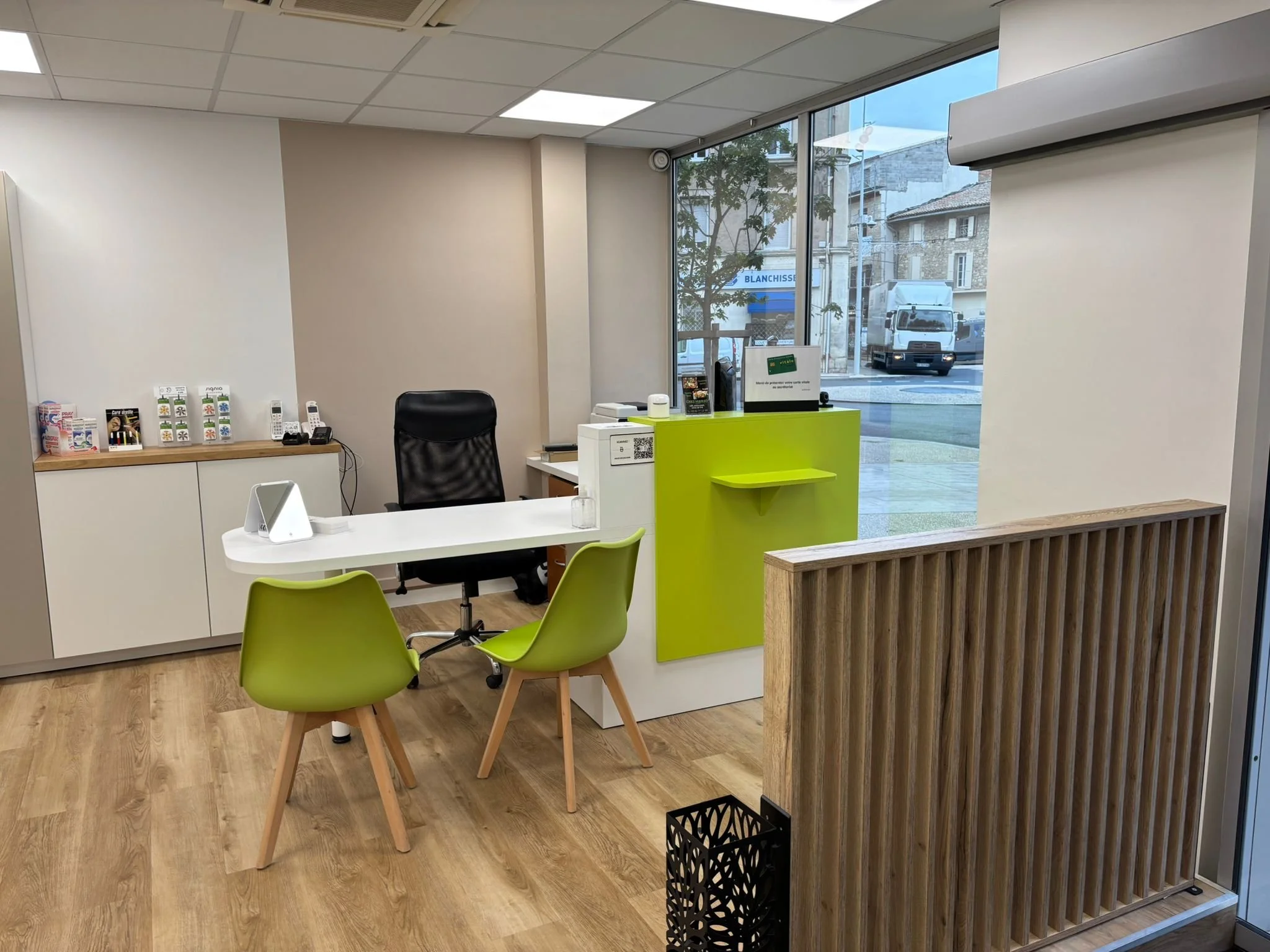 Intérieur d'une réception ou d'un bureau, avec un bureau blanc, deux chaises vertes, un fauteuil noir, une armoires blanche contre le mur, grande fenêtre avec vue sur la rue, sol en bois clair, mobilier moderne.