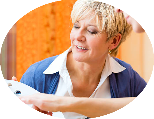 Une femme blonde souriante en blouse bleue, examinant un objet ou un appareil dans un intérieur chaud et lumineux.