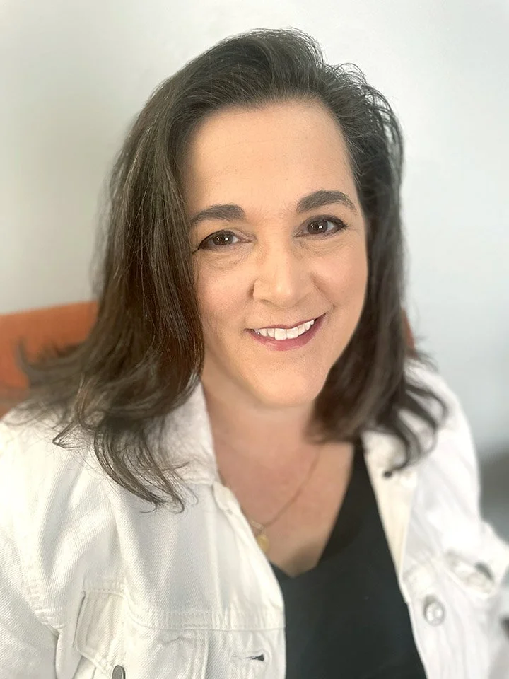 A woman with dark, shoulder-length hair smiling at the camera. She is wearing a white shirt over a black top and a gold necklace, sitting in a well-lit room.