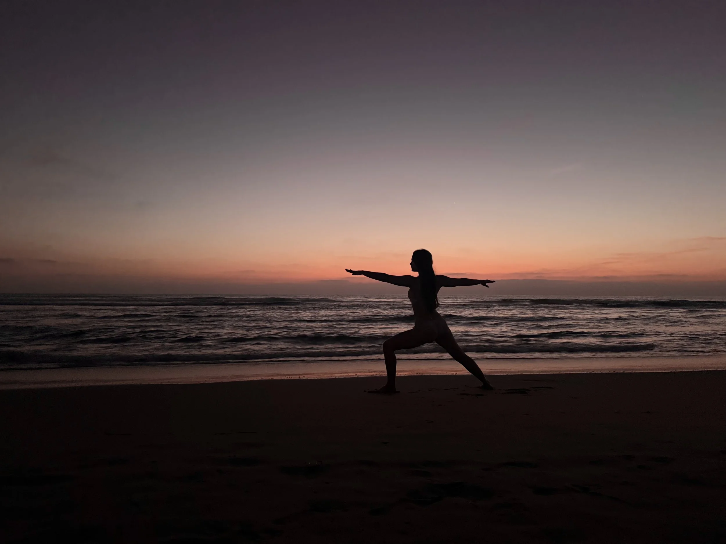 Silhouette of a woman practicing yoga on the beach at sunset with a colorful sky and ocean in the background.
