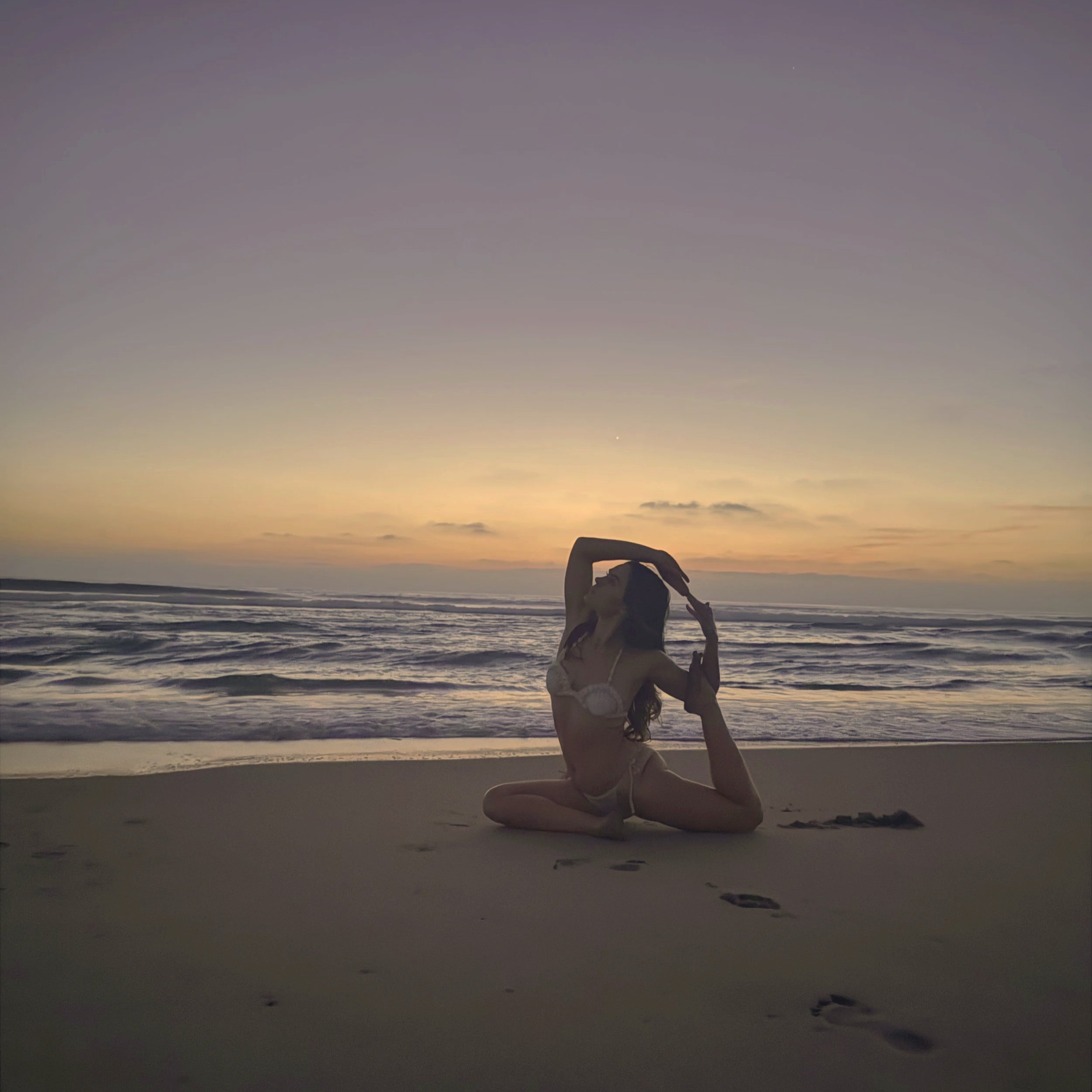 A woman in a bikini practicing yoga on the beach at sunset, sitting in a side stretch pose with her right arm overhead and her left hand on her ankle, overlooking the ocean.