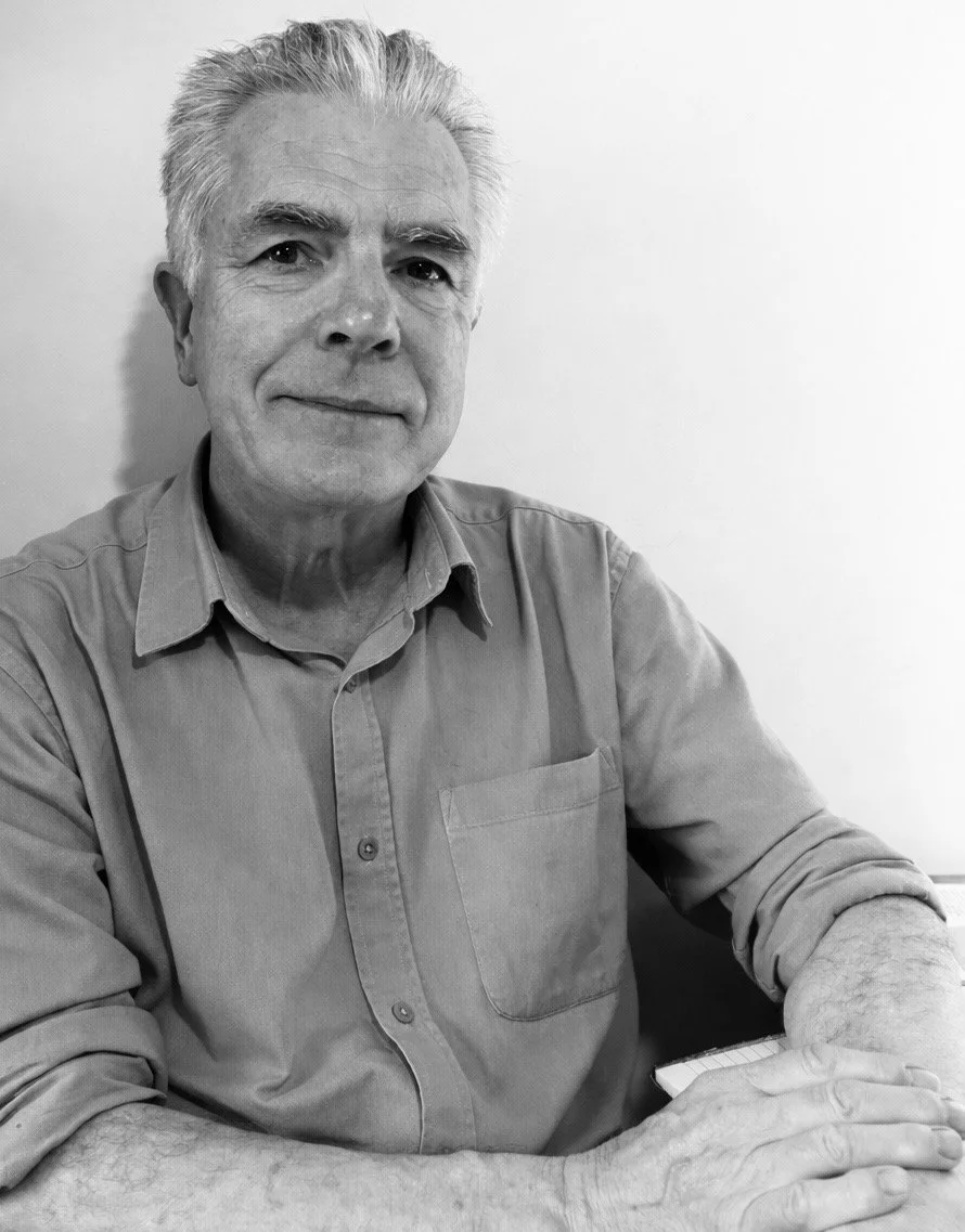 Black and white photo of an older man with gray hair, wearing a button-up shirt, sitting at a table against a plain white wall, smiling softly at the camera.