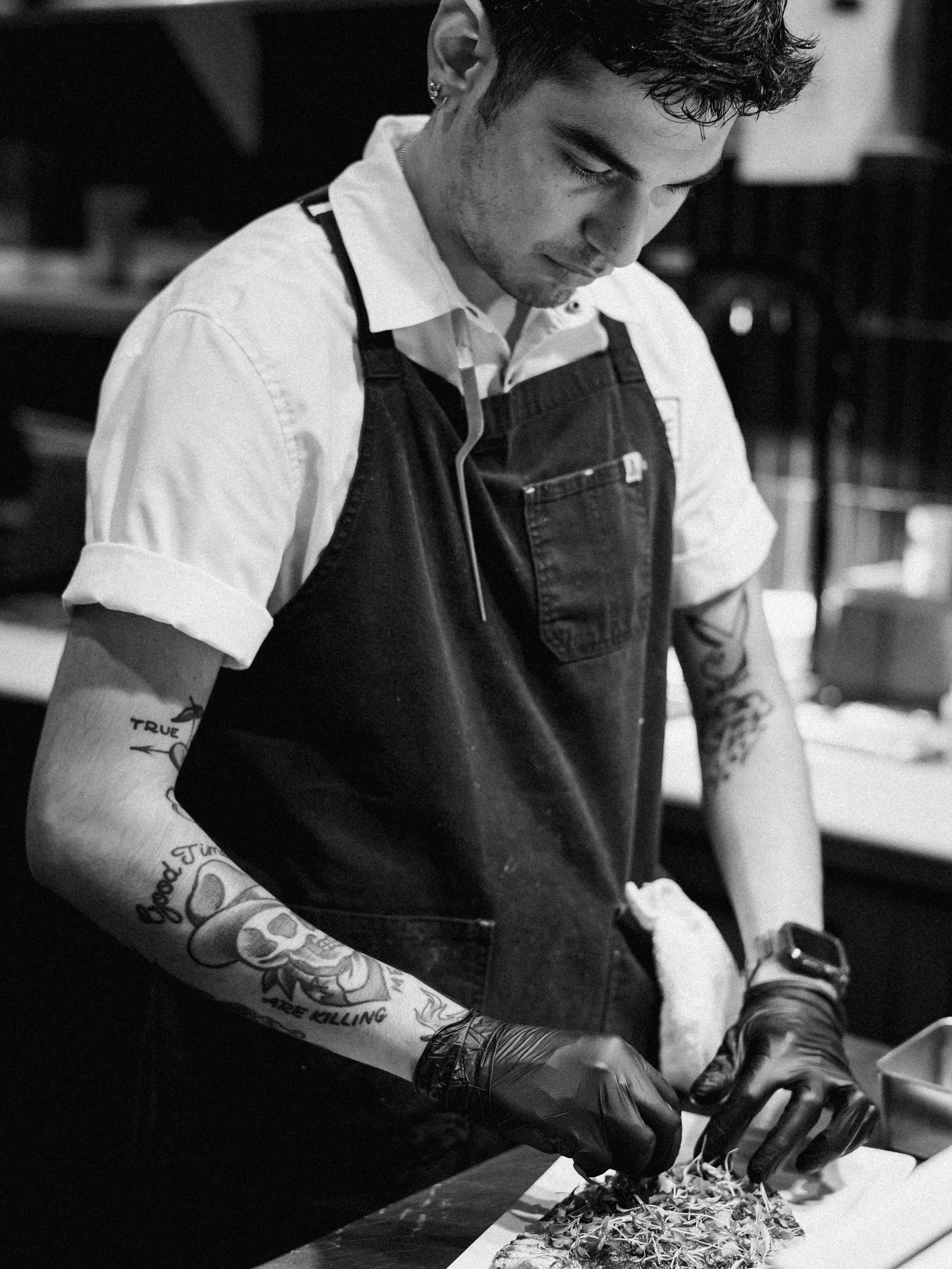 A man with tattoos, wearing a black apron and gloves, preparing food in a kitchen.