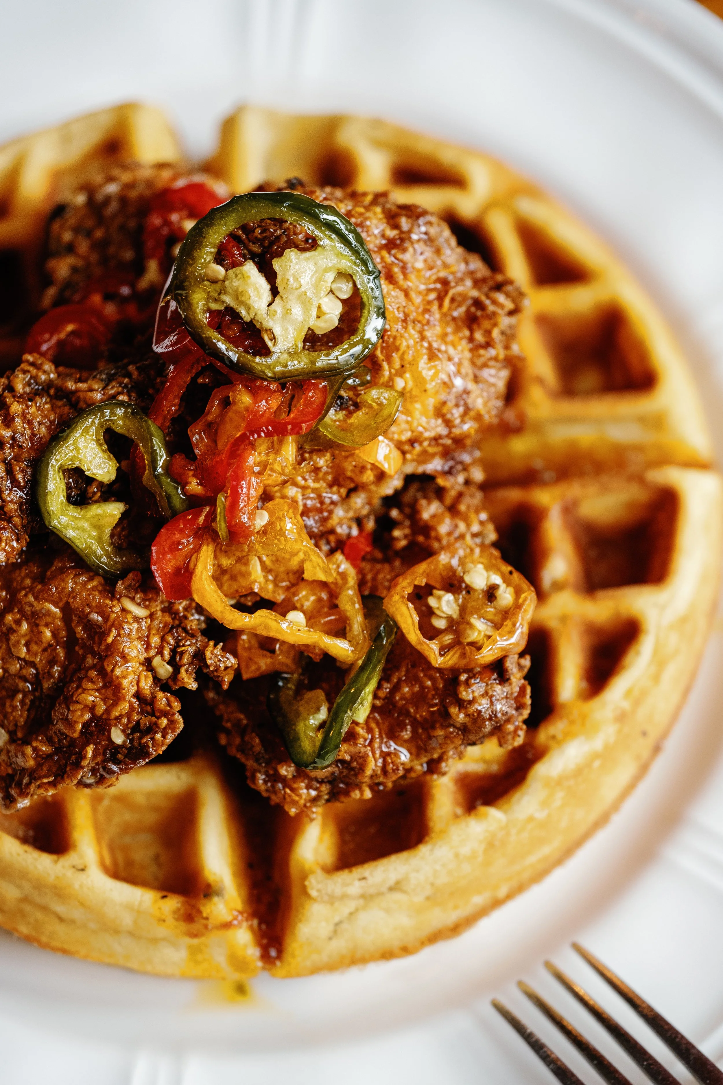 Close-up of chicken and waffles topped with sliced jalapeños and peppers on a white plate with a fork.