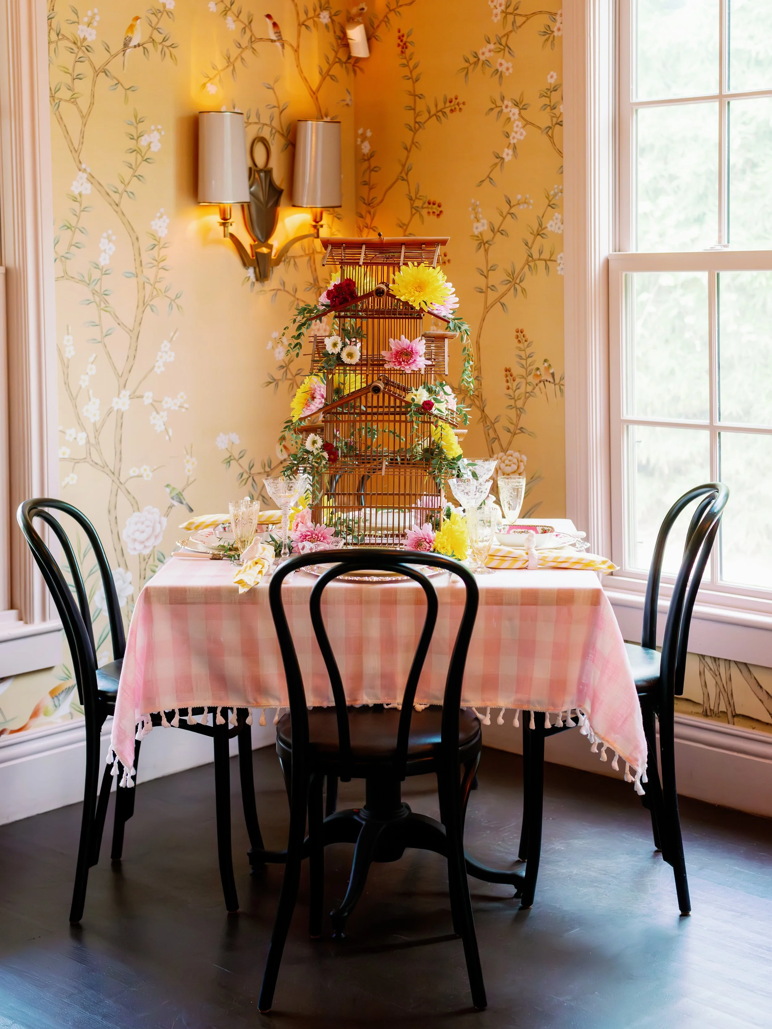 A round dining table with a pink checkered tablecloth surrounded by black chairs, set for a meal with glasses and napkins. The table features a decorative birdcage centerpiece adorned with colorful flowers. The room has large windows, floral wallpaper, and a wall-mounted light fixture.