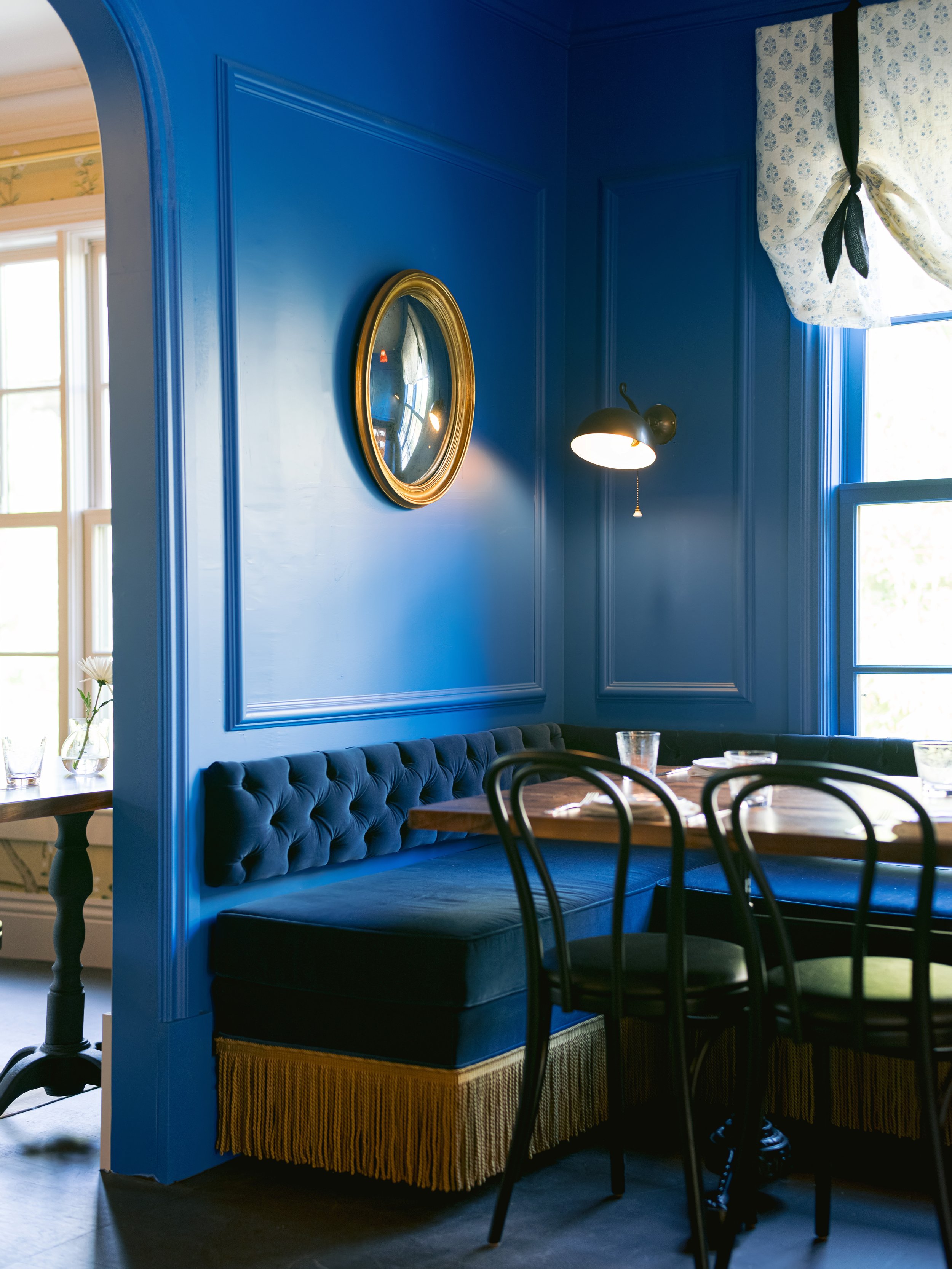 A cozy dining nook with blue walls, tufted blue banquette, and black chairs around a wooden table, near a window with patterned curtains.