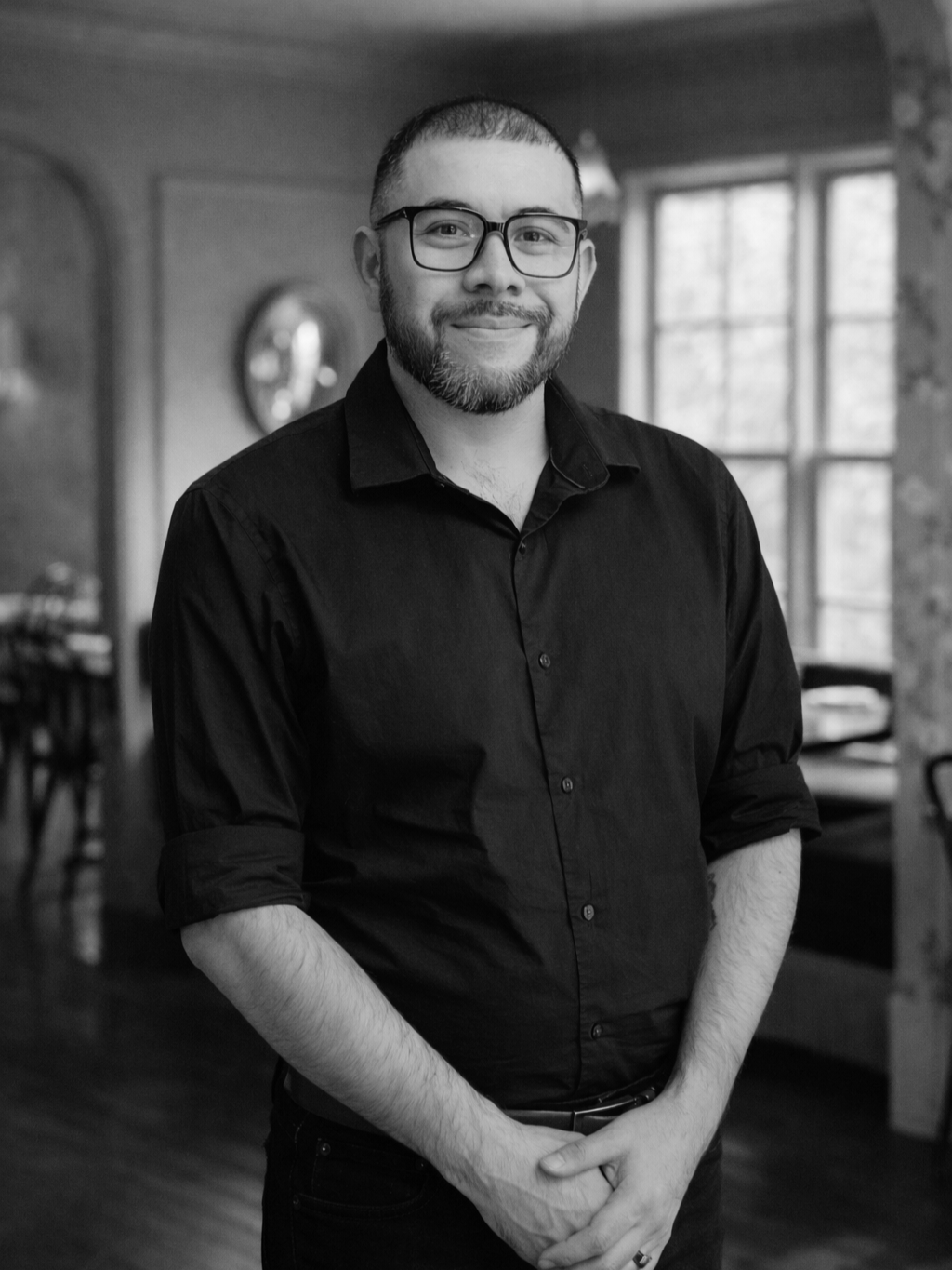 A man with glasses and a beard wearing a black button-up shirt, smiling, standing in a room with large windows and vintage decor.