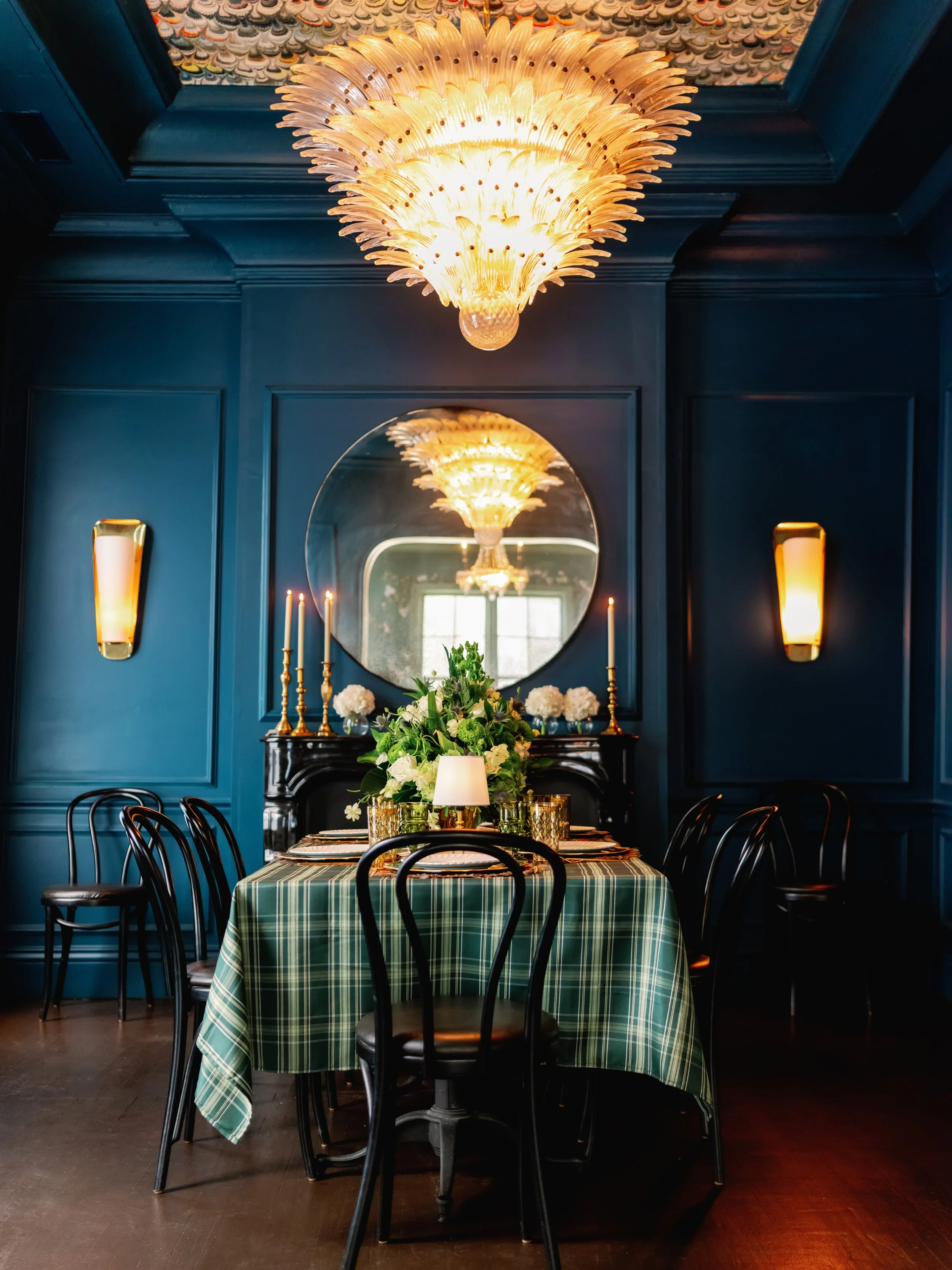 Elegant dining room with a round table covered in a green plaid tablecloth, surrounded by black chairs. The table has floral centerpieces and candles. A large circular mirror with a chandelier reflection and a dark wall with wall sconces complete the setting.