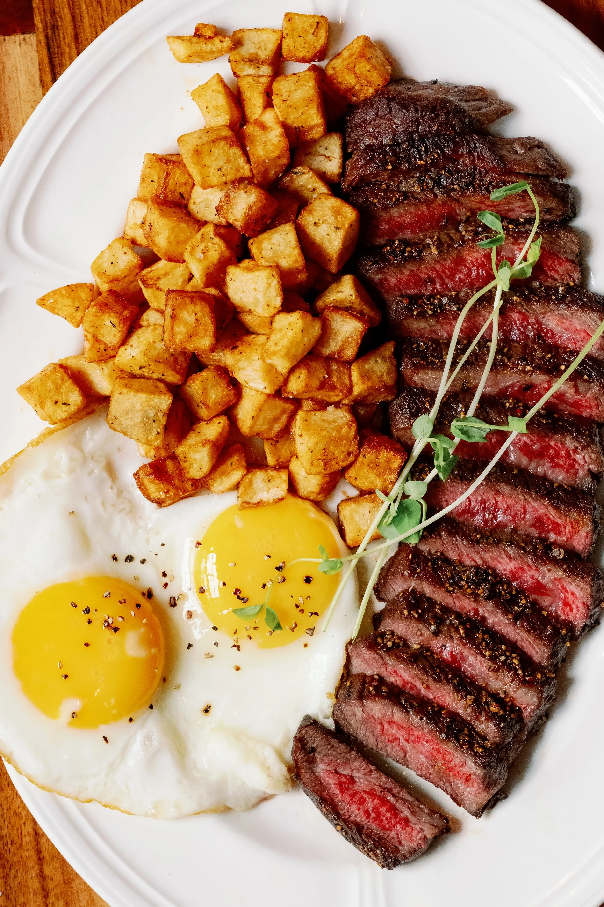 Steak sliced on a white plate with crispy potatoes and sunny side up eggs, garnished with microgreens.