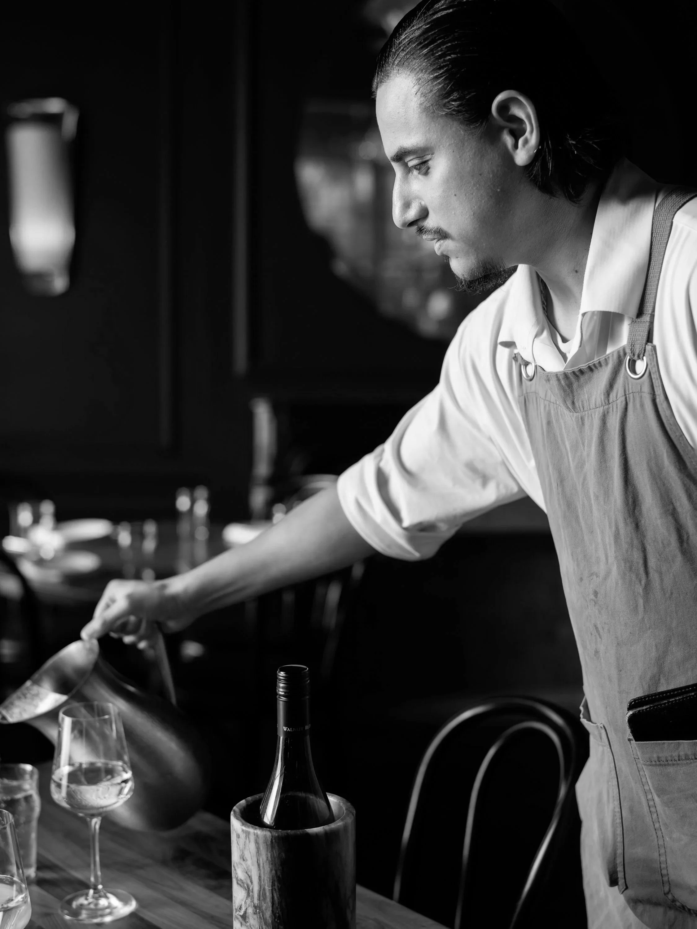 A man pouring wine into a glass on a wooden table, dressed in a light-colored shirt and apron, in a black and white photo.