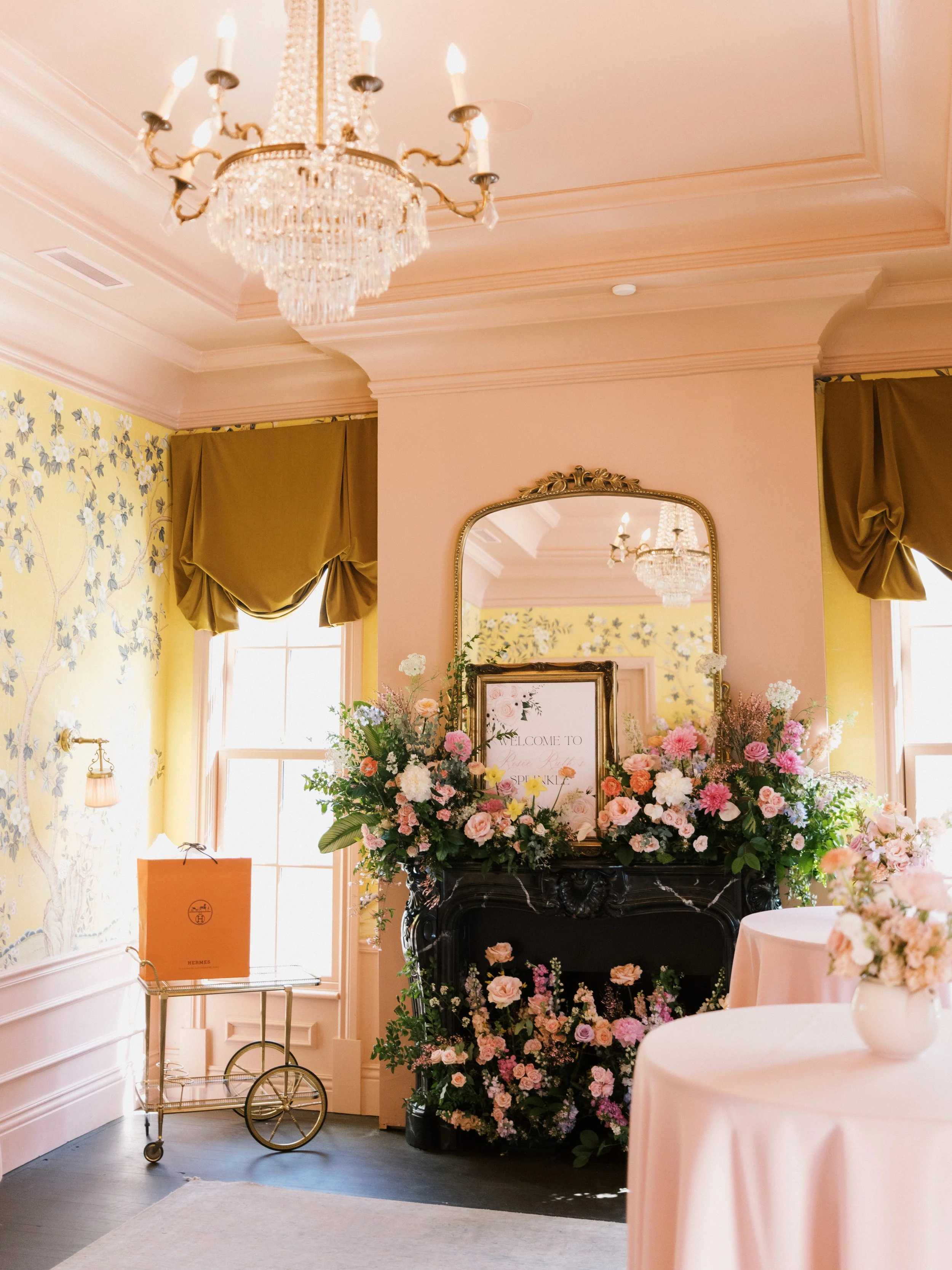 Elegant interior with pink and yellow floral decor, a chandelier, gold curtain drapes, and a decorative black fireplace adorned with pink and white flowers, a framed sign, and a large mirror.