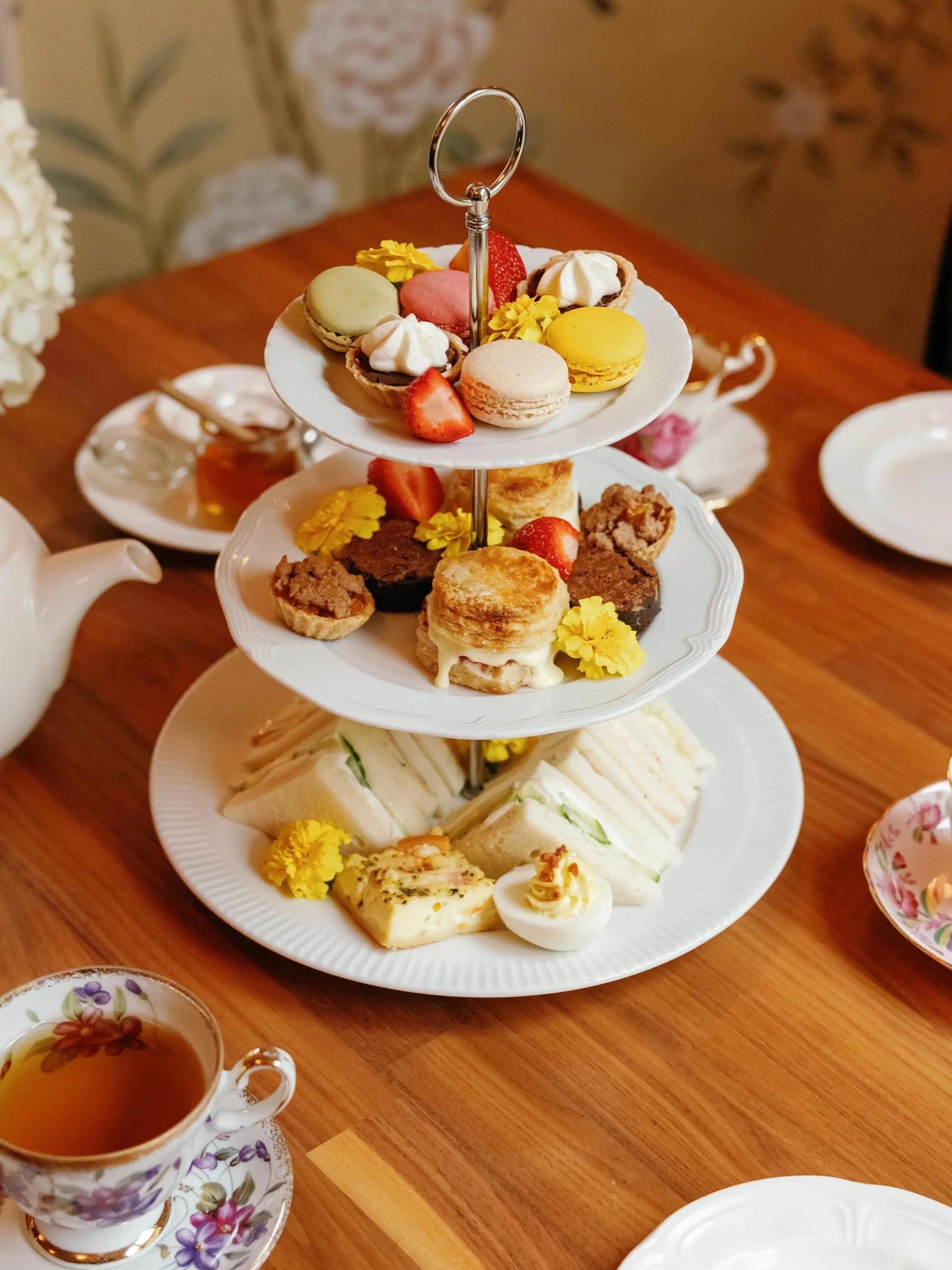Three-tiered serving tray with assorted desserts, finger sandwiches, and flowers, surrounded by tea cups and saucers on a wooden table.